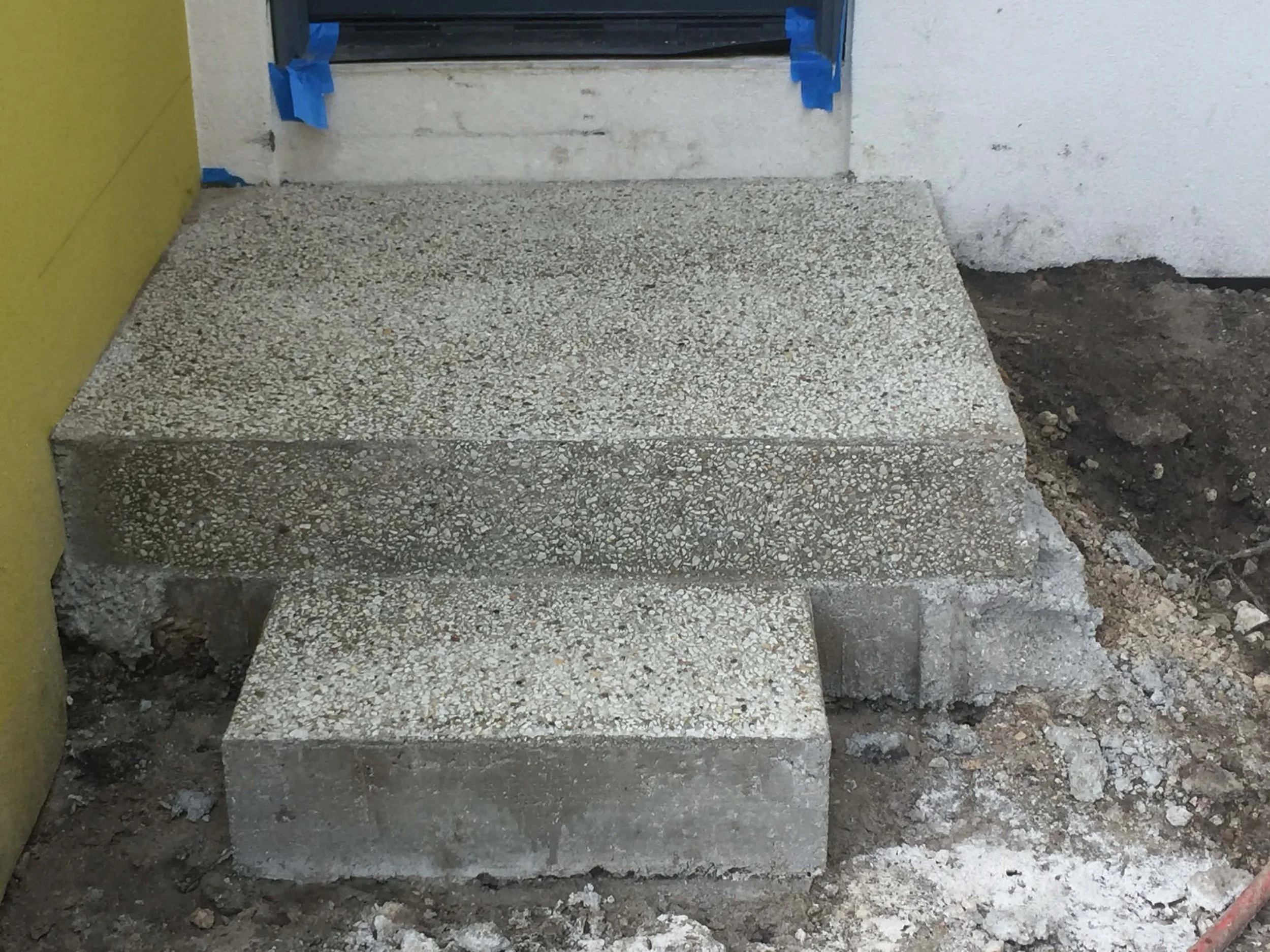 Construction site with concrete steps leading to a doorway, blue tape around the doorway frame, and exposed dirt around the steps.