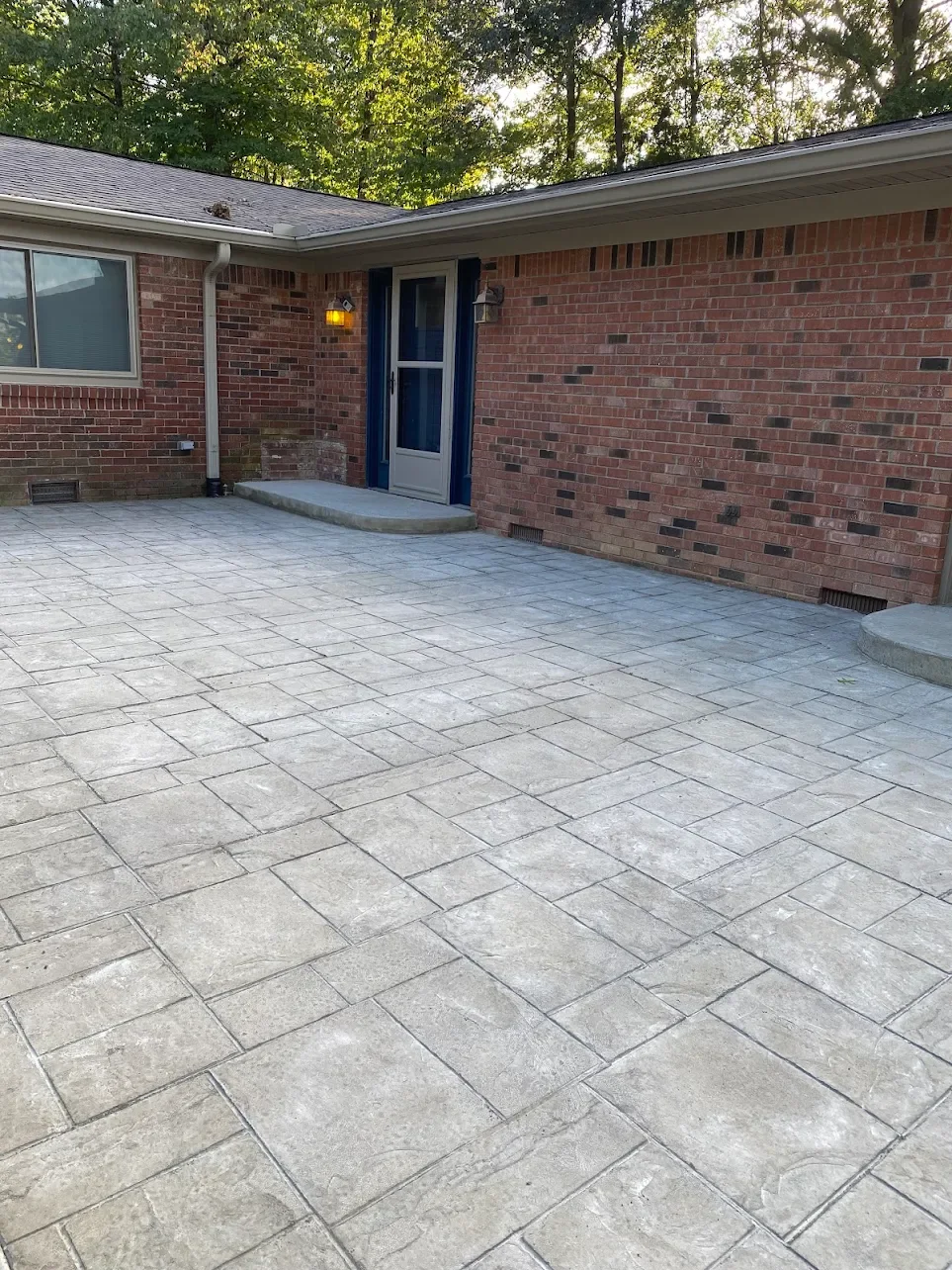 Clean brick house back porch with stamped concrete patio, blue front door with glass panel, sidelight, and outdoor wall lights, surrounded by trees.