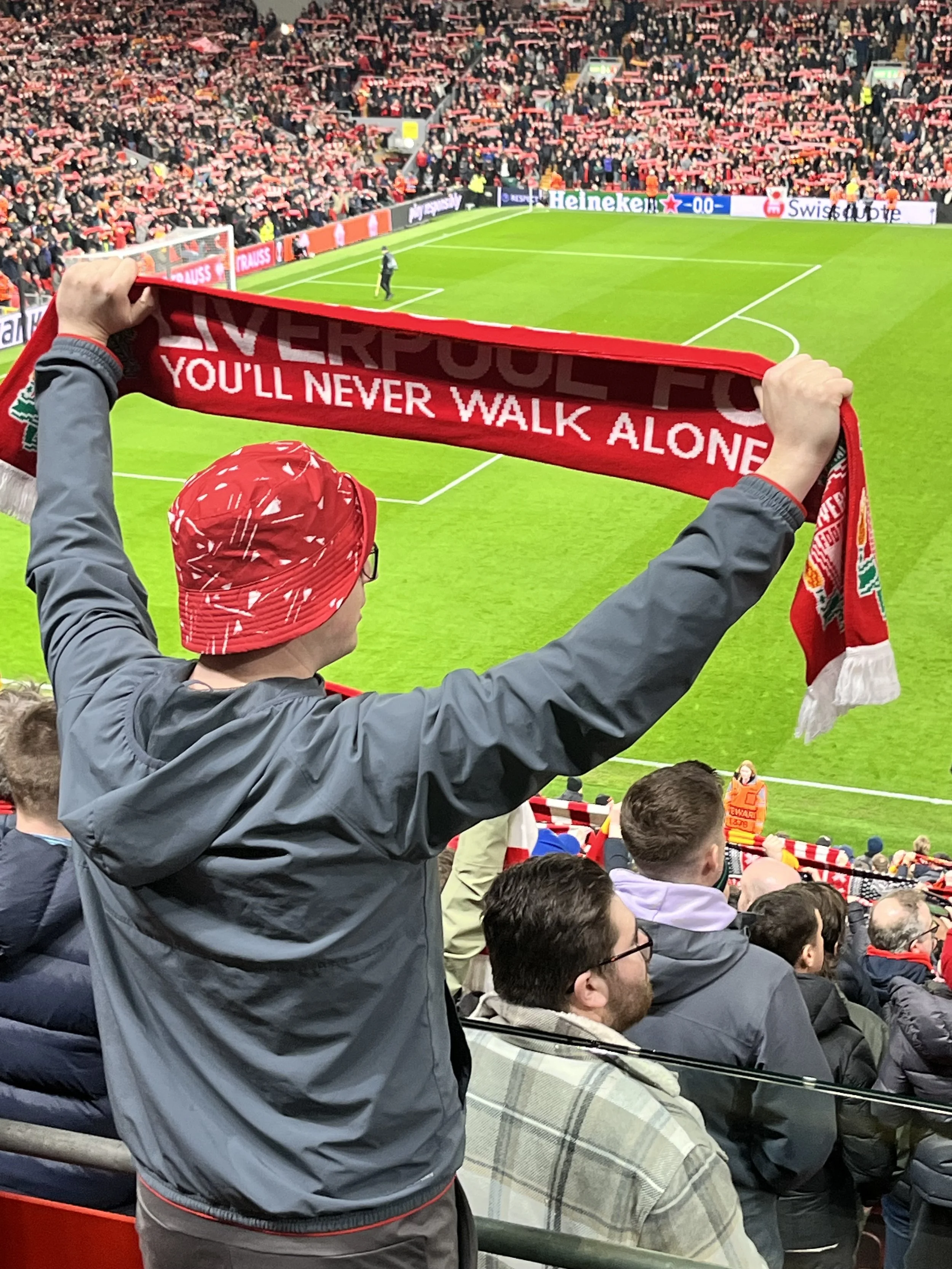 Liverpool FC fan Holing a scarf in a crowded Anfield Stadium. It Reads: You'll Never Walk Alone
