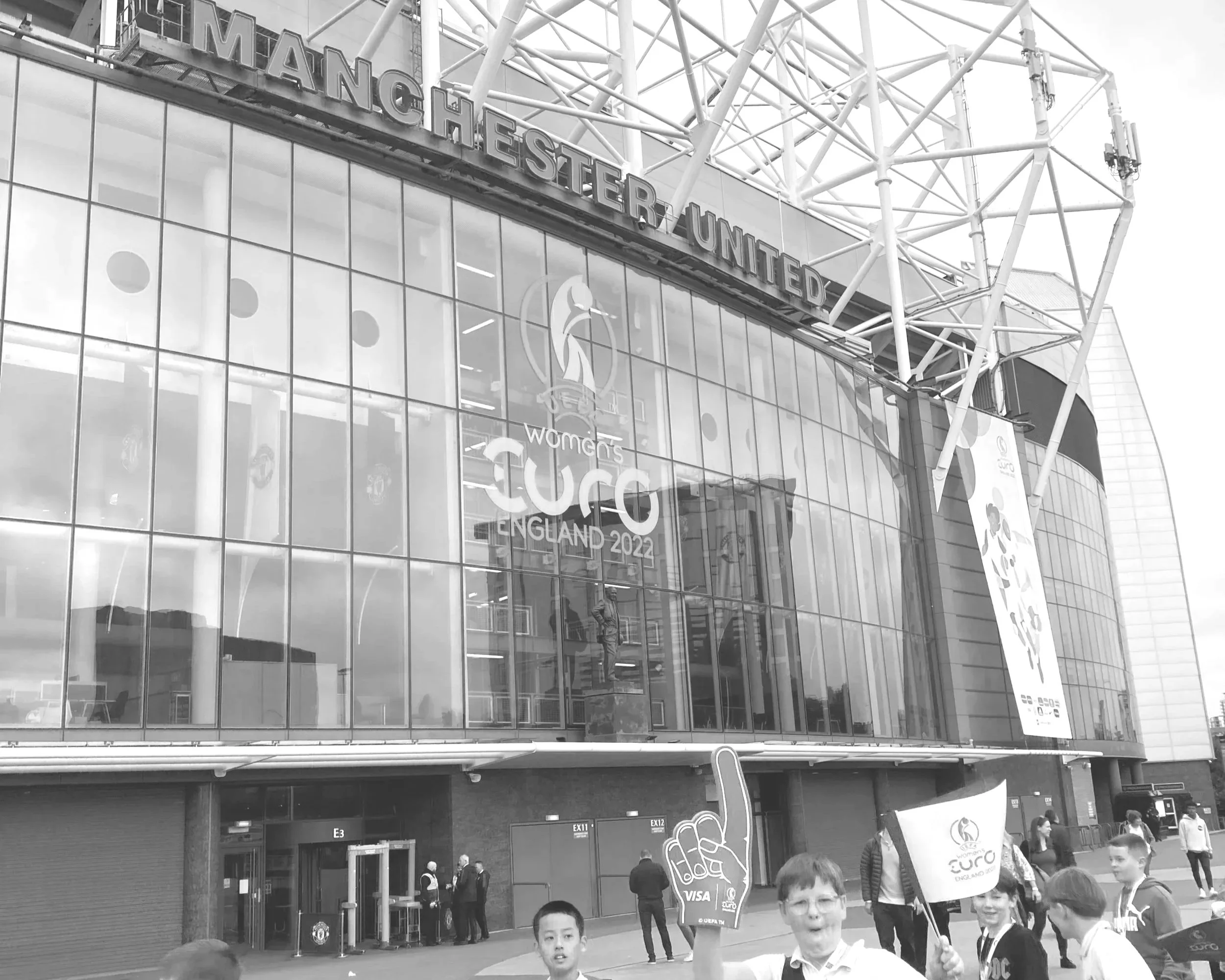 Exterior of the Manchester United stadium with large glass windows and a crowd of young people holding Euro 2022 flags and a foam finger, in front.ⓒSamantha Scott/Think ThreeSixty.