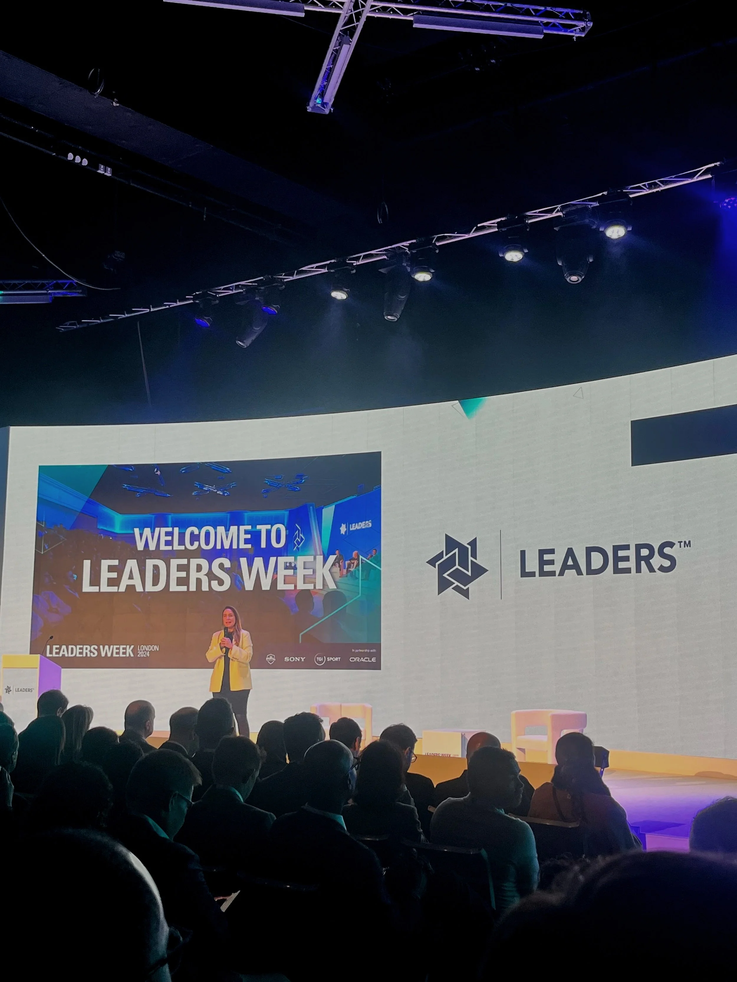  A stage with the crowd looking at the large LED Leaders Week backdrop and female speaker