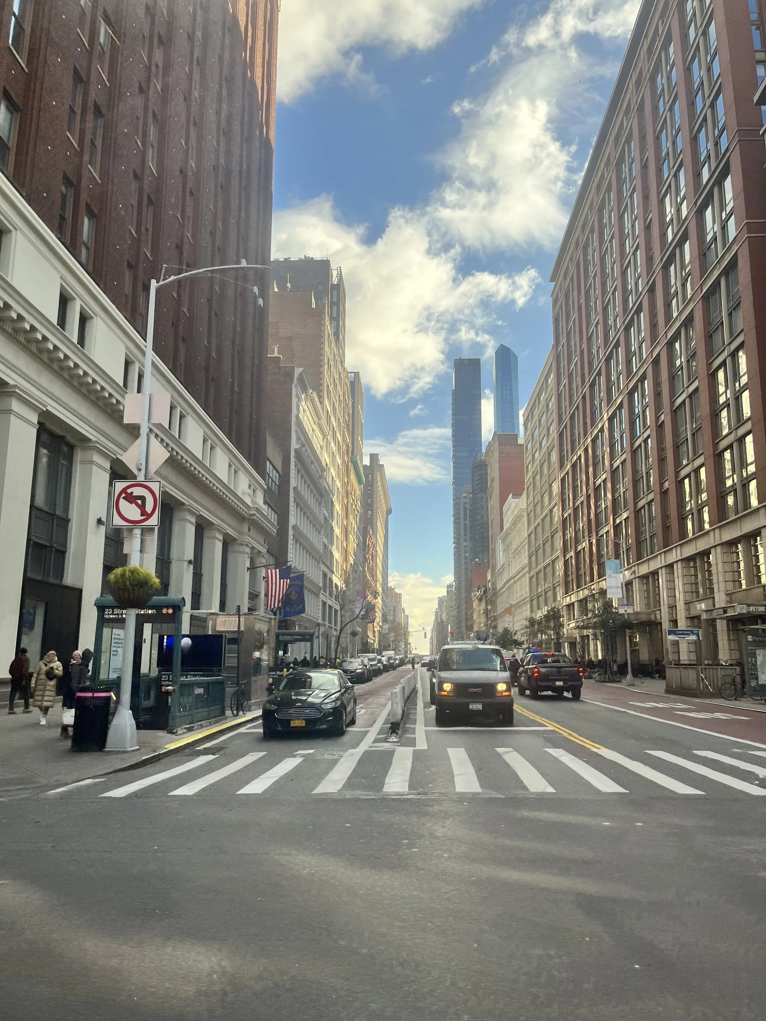 New York street showing high rise buildings and cars at a crosswalk. The sky is bright blue and shows between the buildings