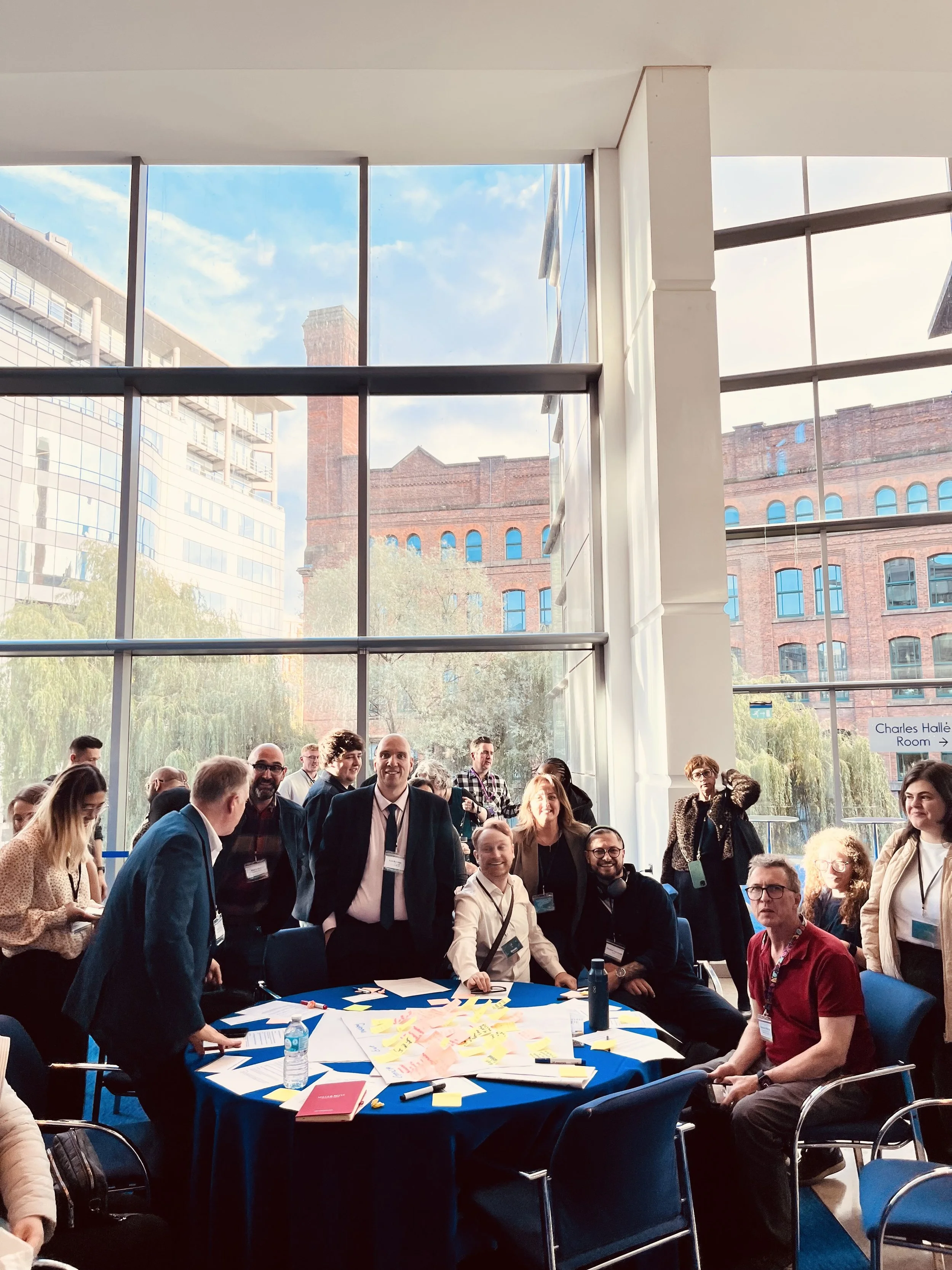 Group image at a GMCA conference tackling the digital divide. there is a large mixed group of around 20 people at a table discussing and change and barriers affecting people across the region