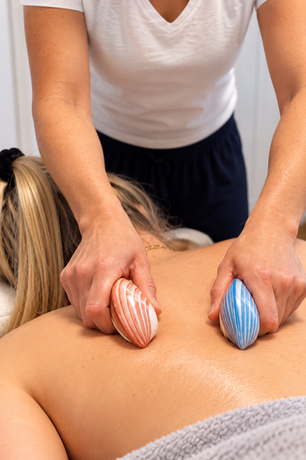A person receiving a massage with two large, colorful stones on their back, one red and cream and the other blue and white, in a spa or massage setting.