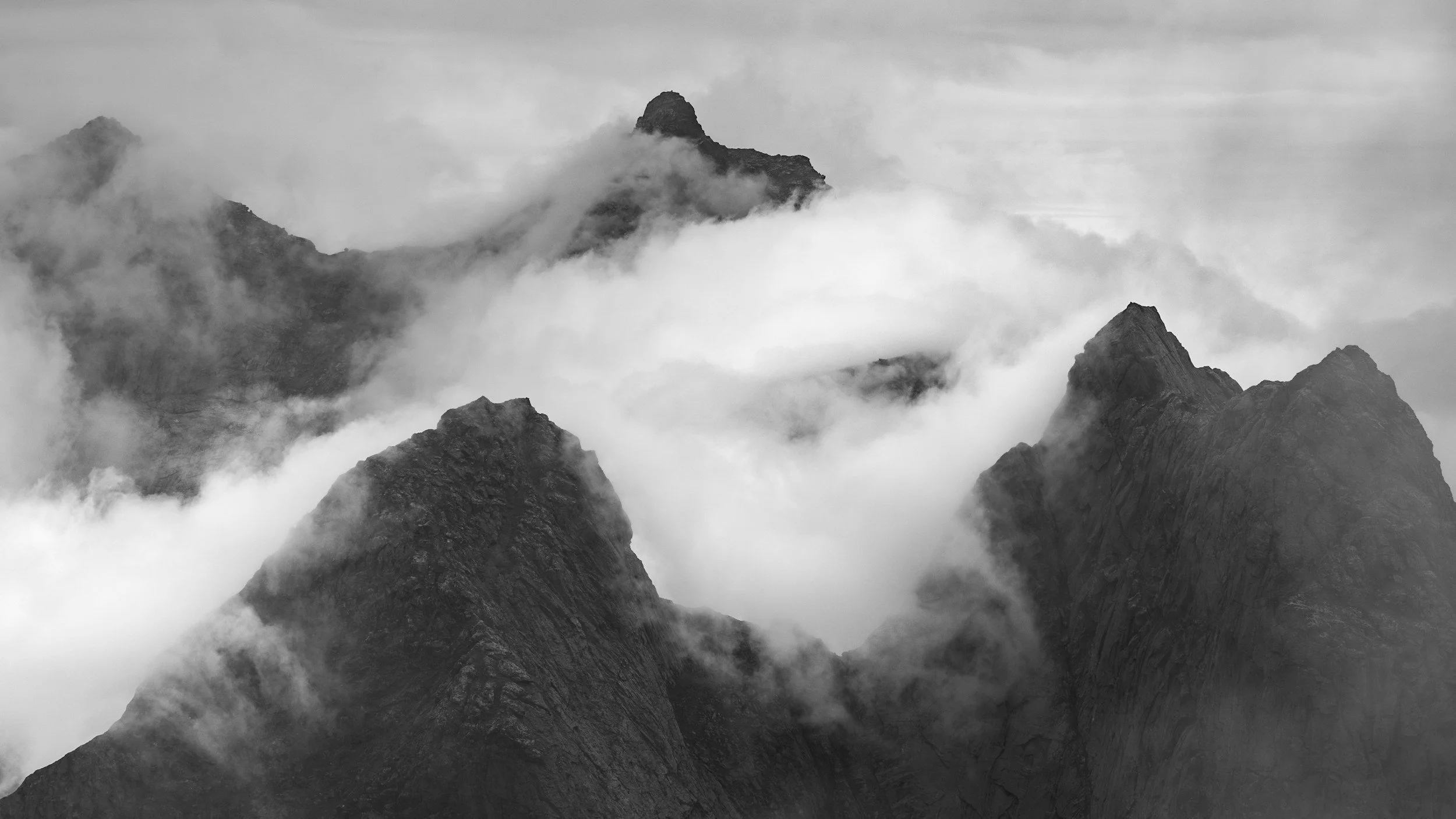 Black and white photograph of rugged mountain peaks shrouded in clouds, creating a misty atmosphere.