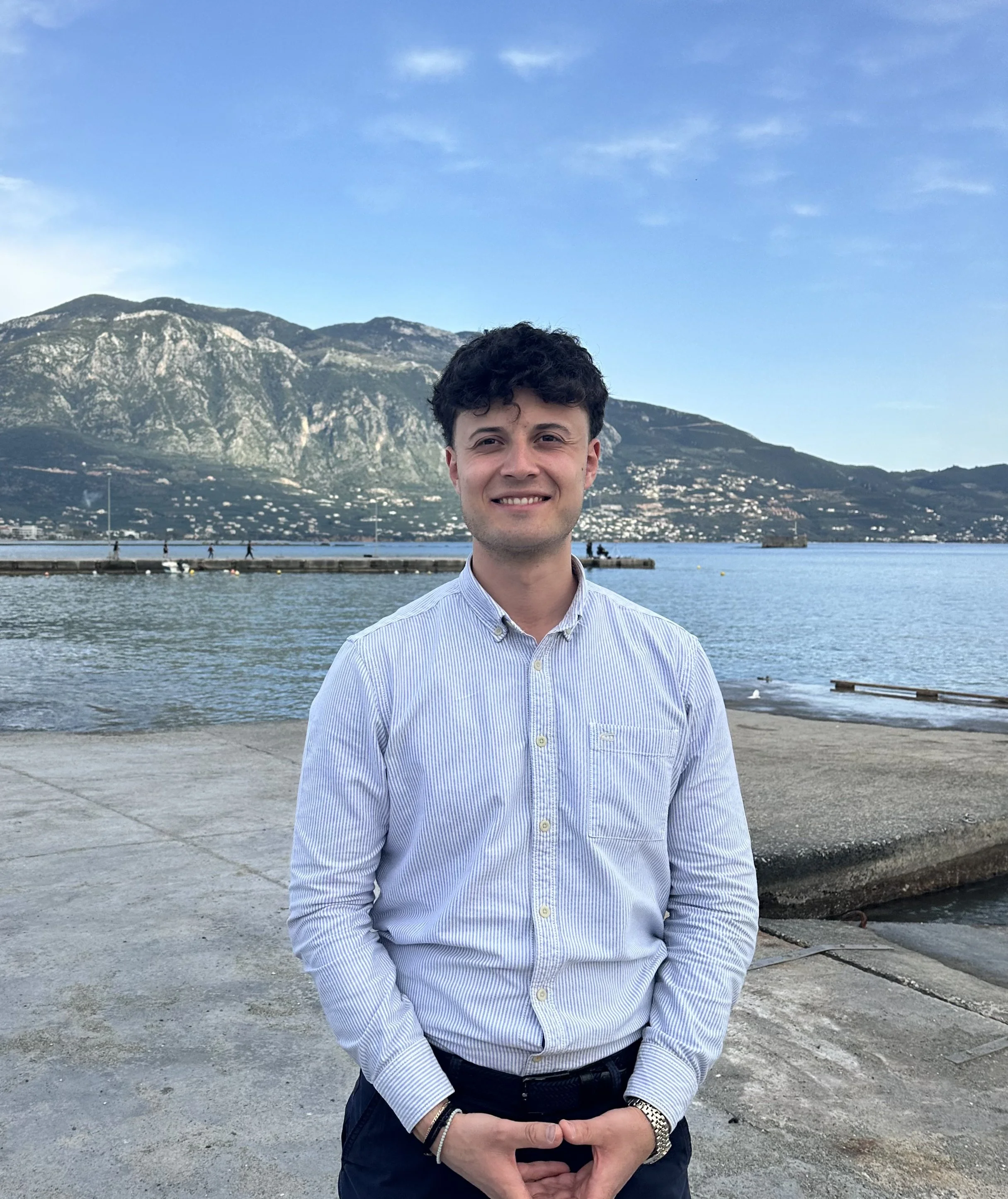 A young man with dark, curly hair smiling while standing outdoors by a body of water with mountains in the background, wearing a light blue, striped button-up shirt and dark pants.