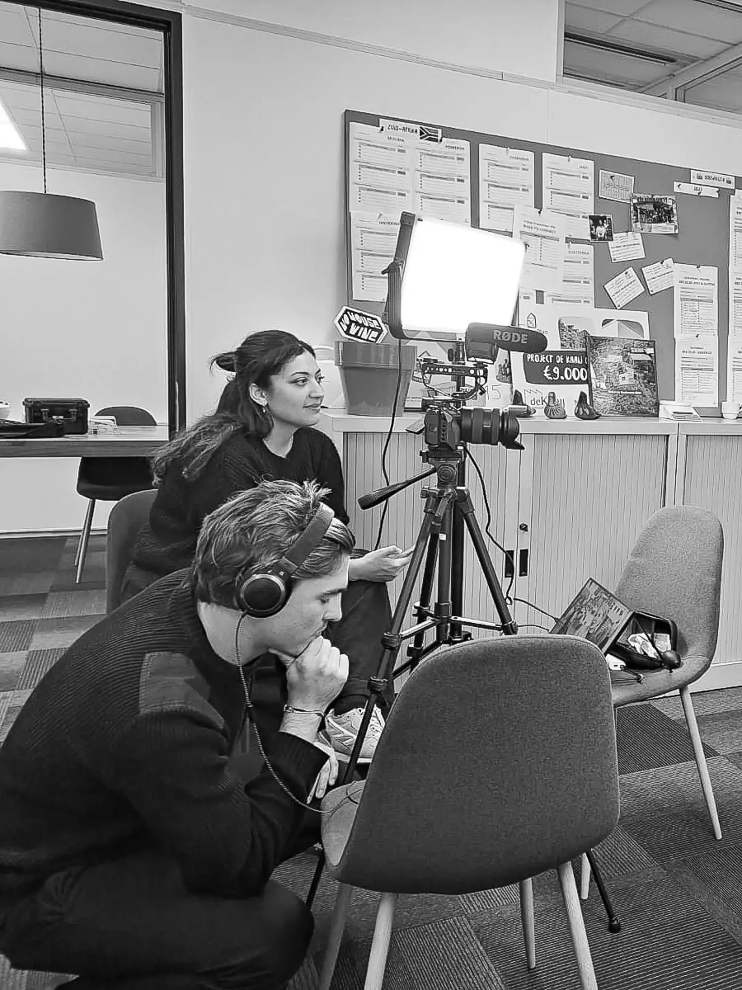 Twee mensen van bedrijf Motion Stream met audiogear, een camera op een statief en lampen op statieven, aan het werk tijdens een interview setting in een kantoorruimte.