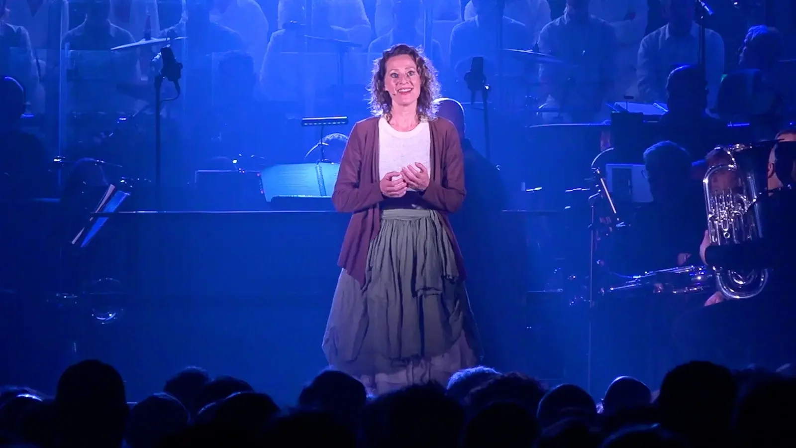 Een vrouw staat op een podium tijdens een musical show, omgeven door een symfonieorkest onder blauwe verlichting. Voor haar zijn wat hoofden te zien van het publiek.
