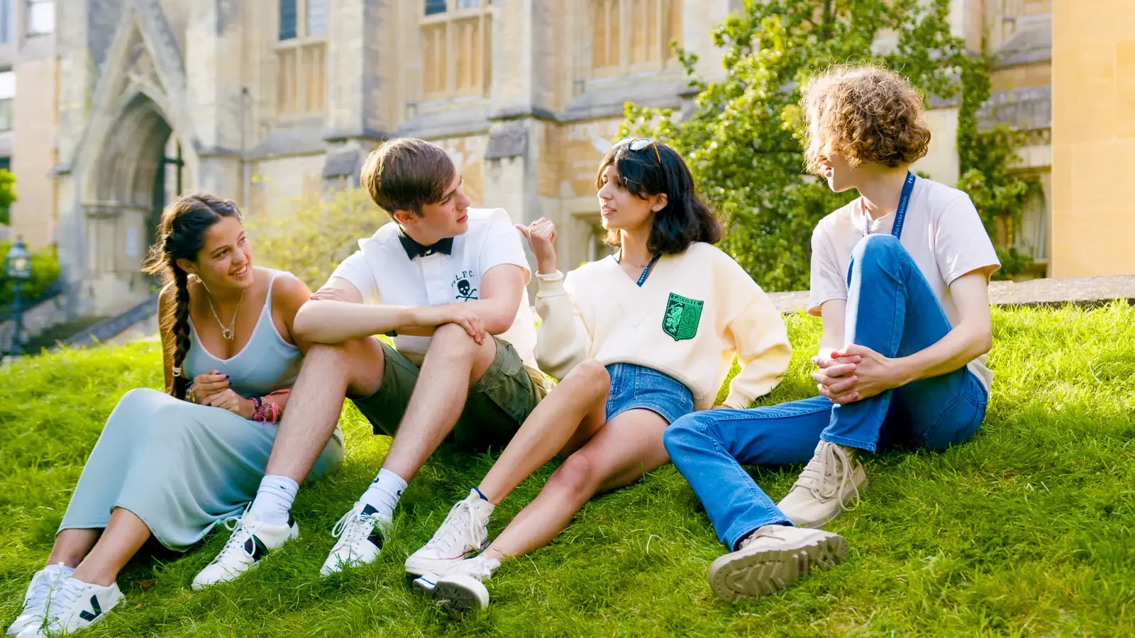 Vijf jongeren zitten op een grasveld voor een oud schoolgebouw, ze praten en lachen tijdens een zonnige dag.