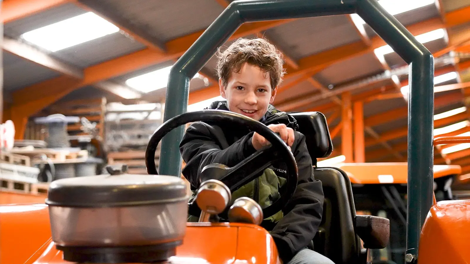 Kind lacht naar de camera terwijl hij op een tractor zit in een magazijn vol met met opslagrekken.