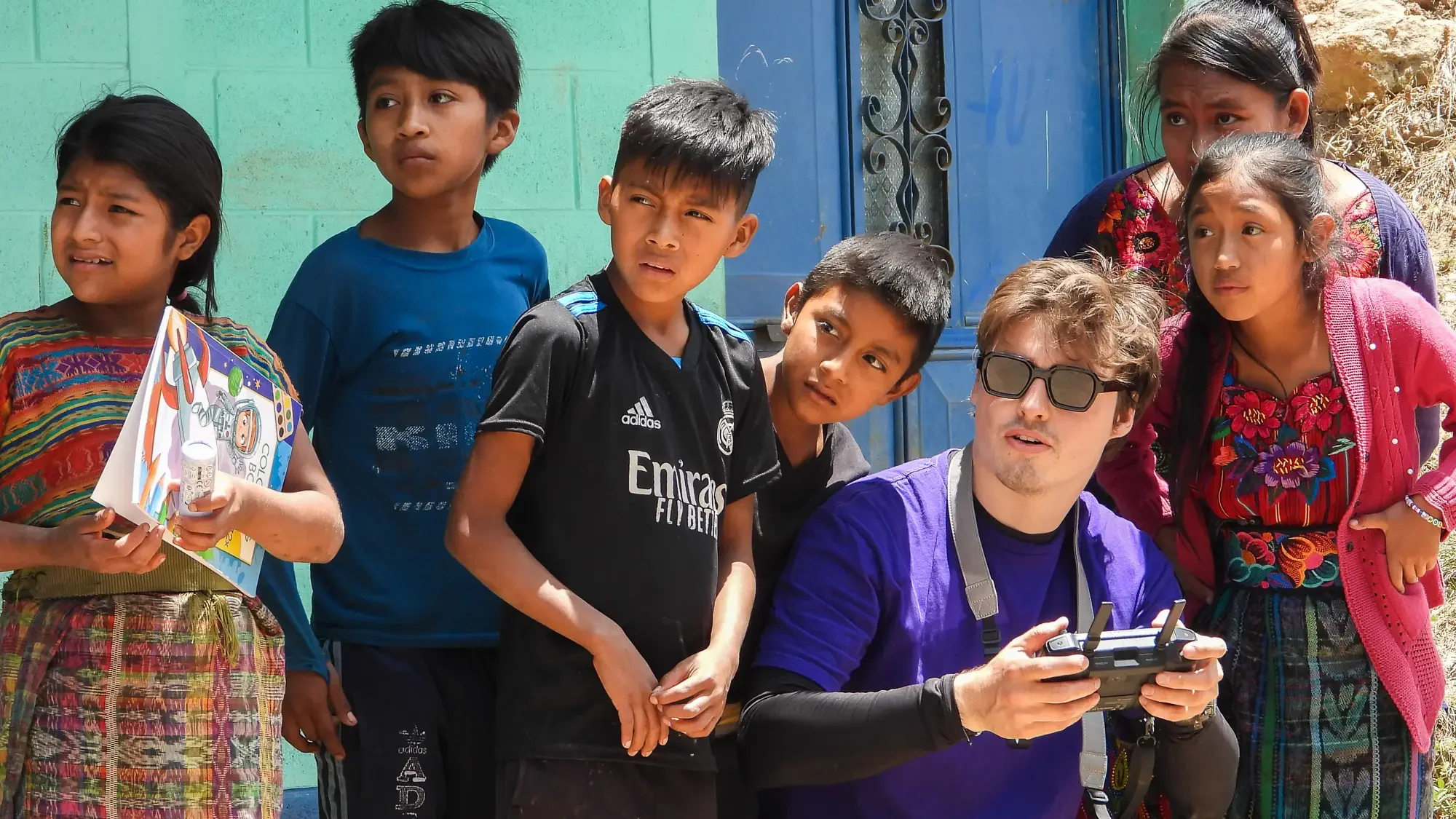 Een man met een drone-controller omringd door zeven jonge kinderen in Guatemala. Het is zonnig en ze staan buiten voor een blauwe deur en een groene muur.