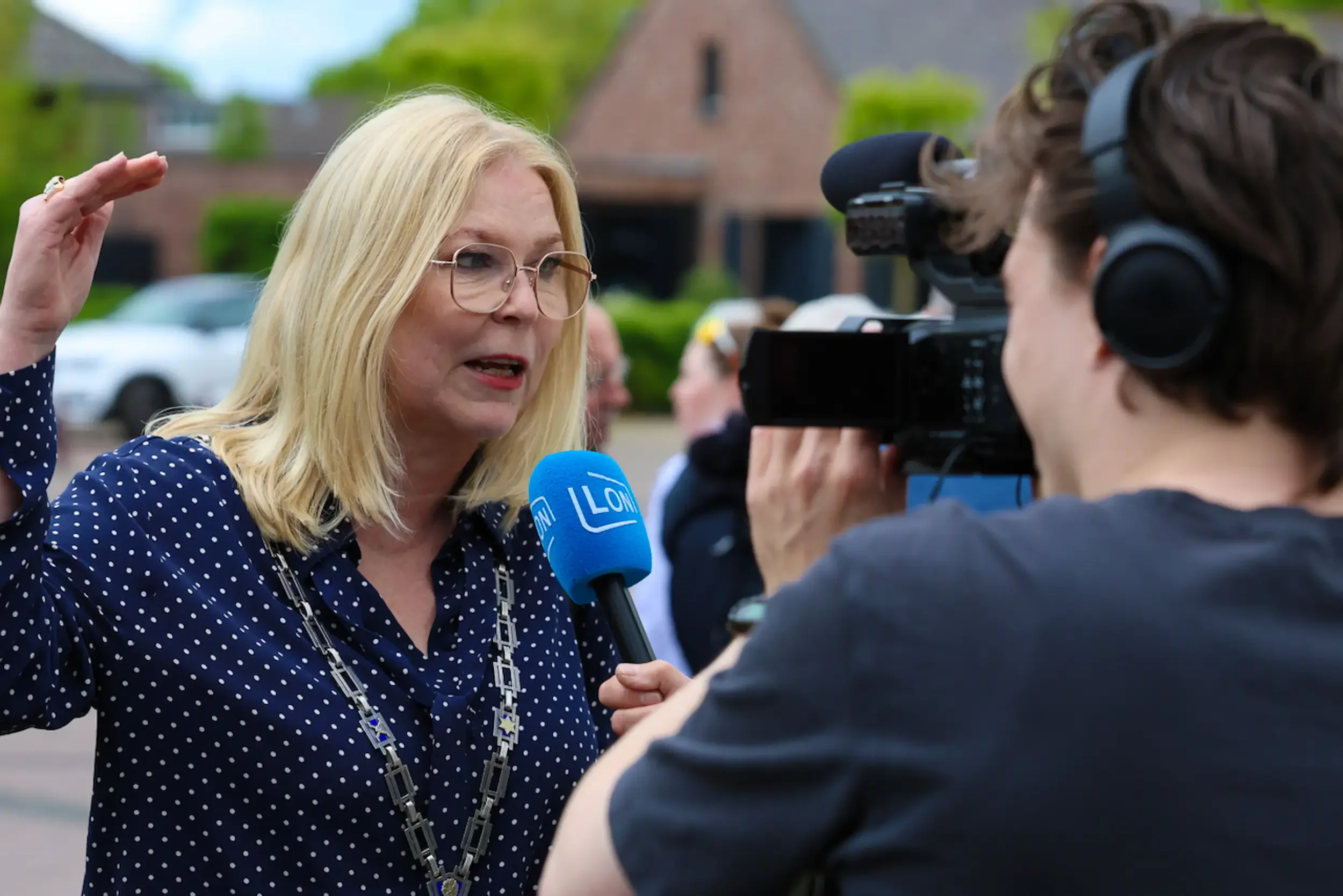 Een vrouw met blond haar, een bril en een polkadot blouse (de voormalige burgemeester van het dorp Nuenen) wordt geïnterviewd door een journalist met een camera en microfoon tijdens een buitengebeurtenis. Op de microfoon staat de tekst "LON".