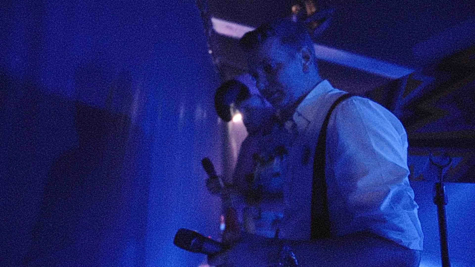 Twee mensen met allebei microfoons in hun handen een donkere ruimte met blauwe verlichting. Ze staan voor een groot doek, backstage van een concert.