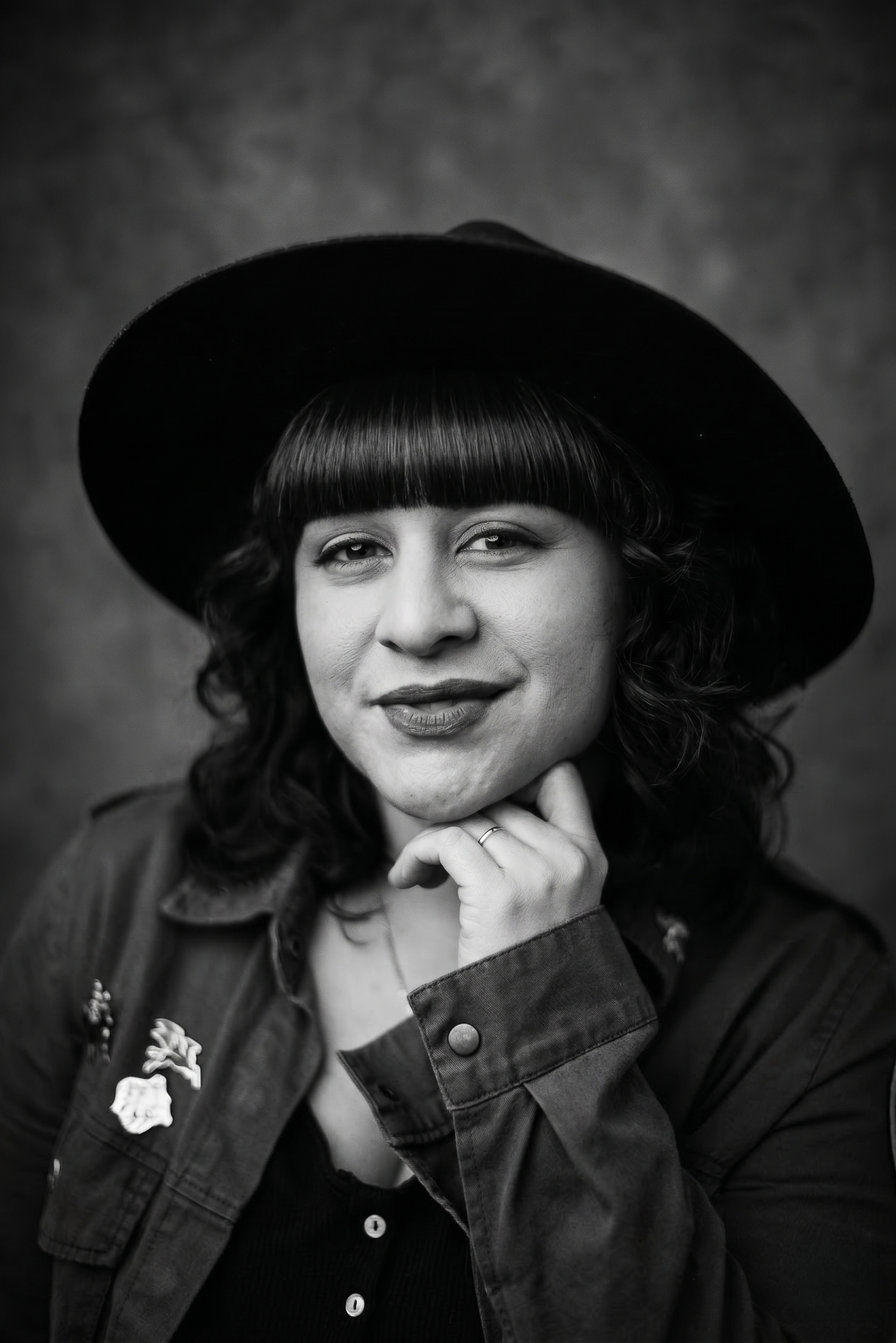 Black and white portrait of a Kyleene Finley wearing a wide-brimmed hat and a denim jacket, resting her chin on her hand, with a slight smile and curly hair.