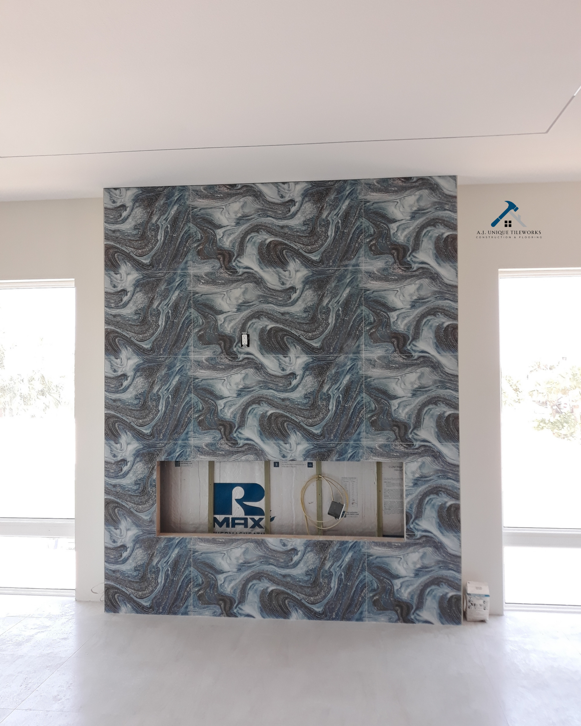 Interior of a room with a decorative marble-patterned wall with a rectangular cutout, adjacent to two windows, construction materials inside the cutout, and a logo for 'A.J. Unique Tileworks' in the top right corner.