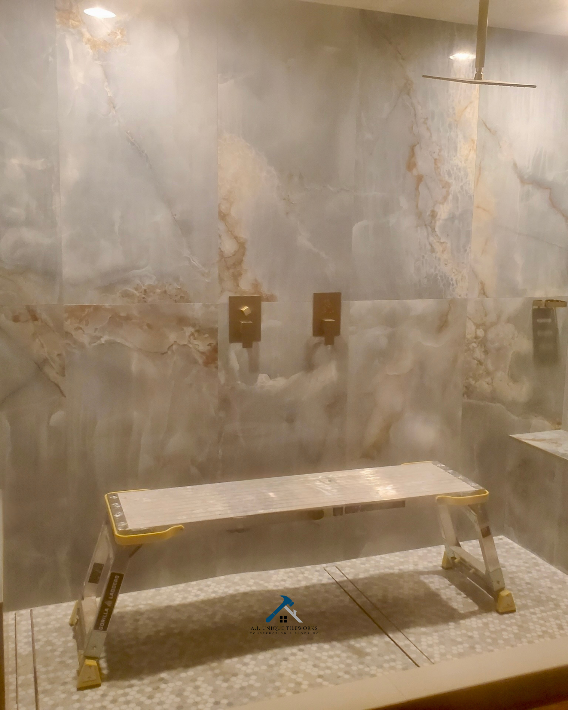 Shower area with marble walls, a showerhead, and a bench under construction, with a step-in area. Gold fixtures and a textured floor.
