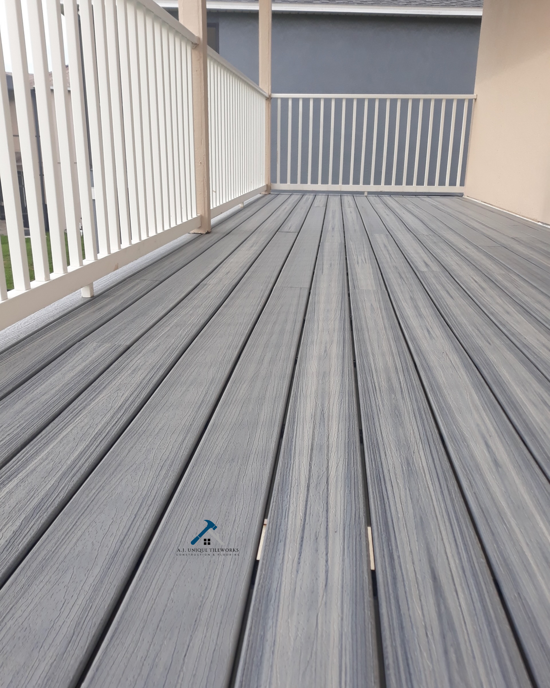 An outdoor balcony with wooden gray composite decking and white railings, with a small logo of a hammer and words "A.J. Unique Tileworks."