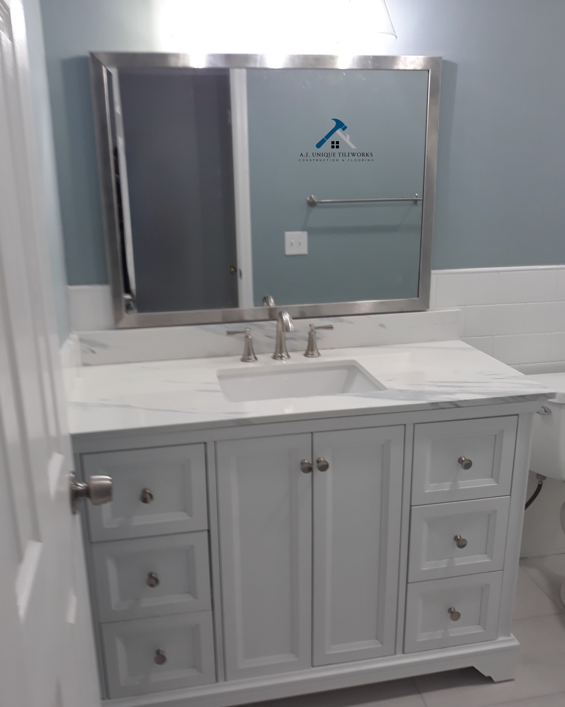 White bathroom vanity with drawers and cabinet doors, marble countertop, and undermount sink in front of a large mirror with a logo reading 'A.J. Unique Tileworks Construction & Flooring' on the mirror.