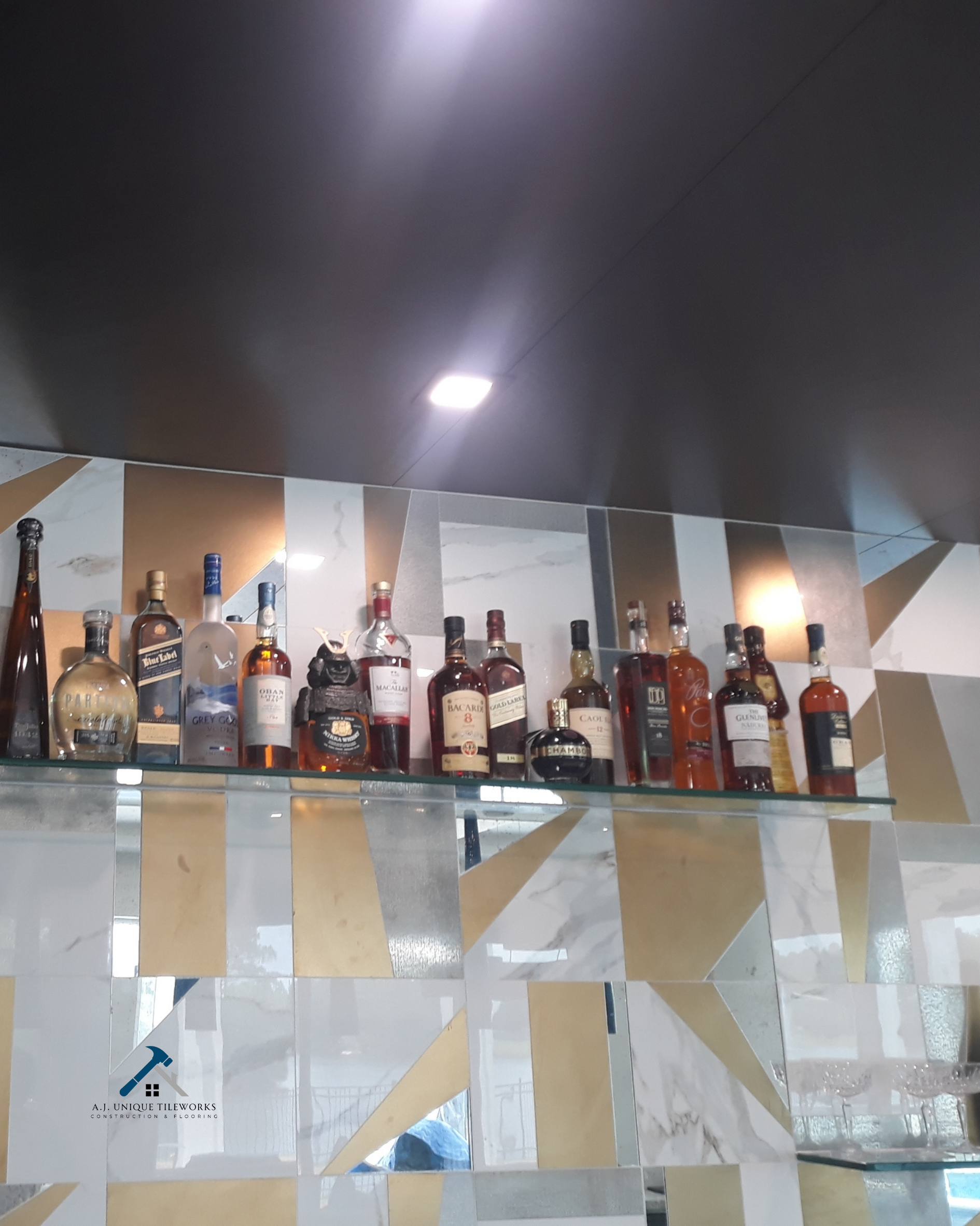 A display of various bottles of liquor on a glass shelf against a geometric patterned tile wall in a bar or lounge area.