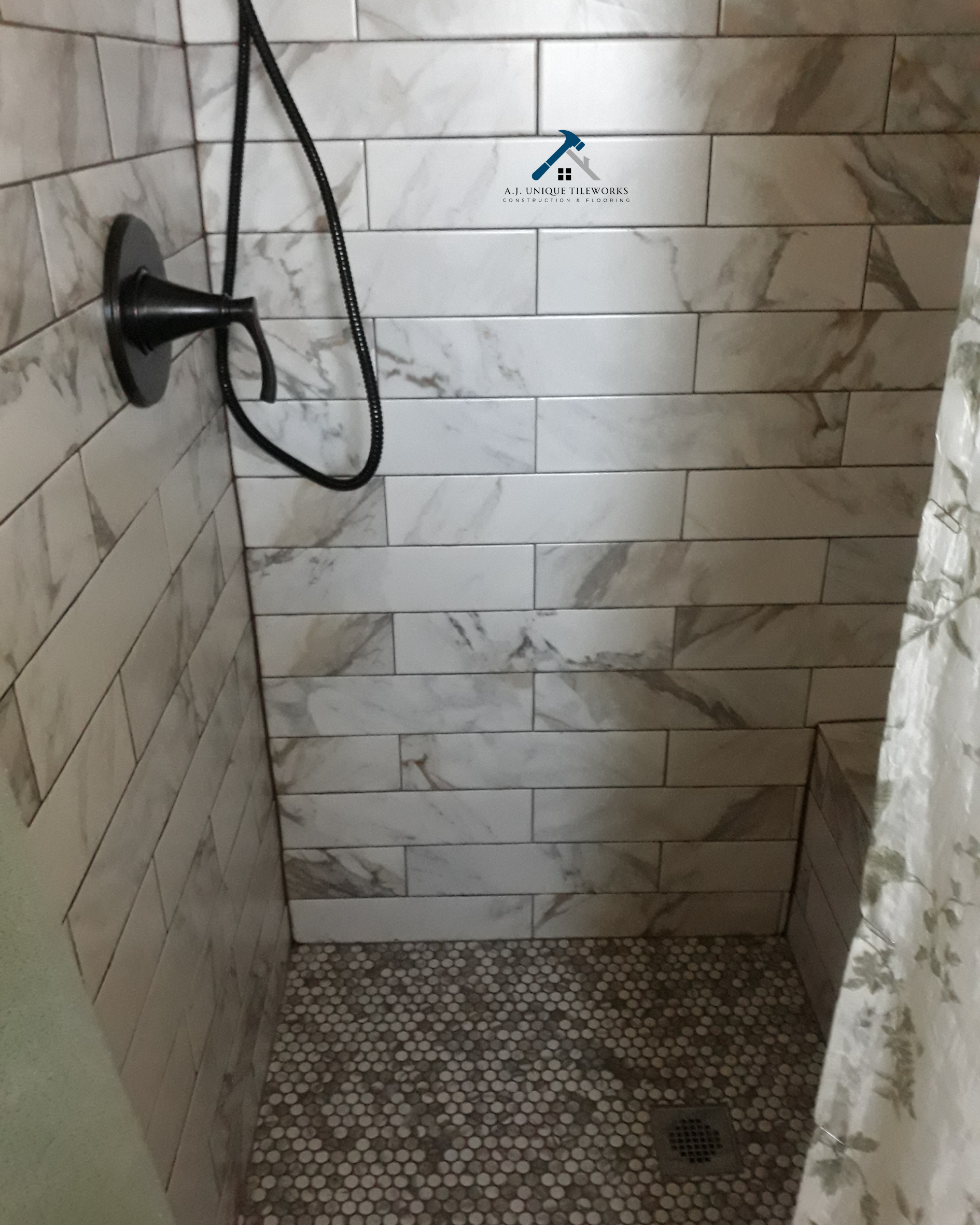 A shower with marble tile walls and a pebble stone floor, featuring a black showerhead and hose, and a drain on the floor.