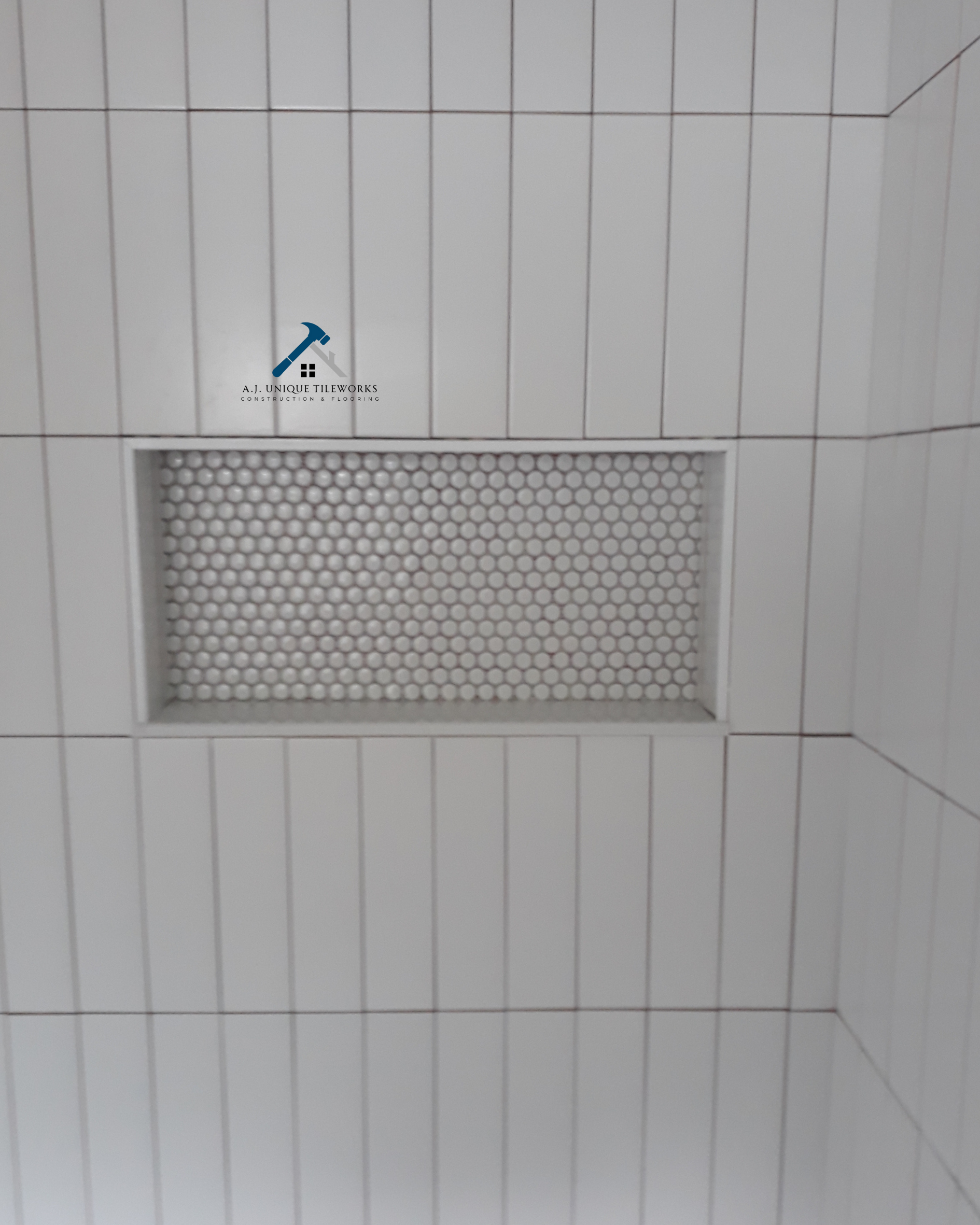 White tiled wall with a rectangular air vent covered by a honeycomb-patterned grille, and a logo of A.J. Unique Tileworks featuring a hammer and house icon.