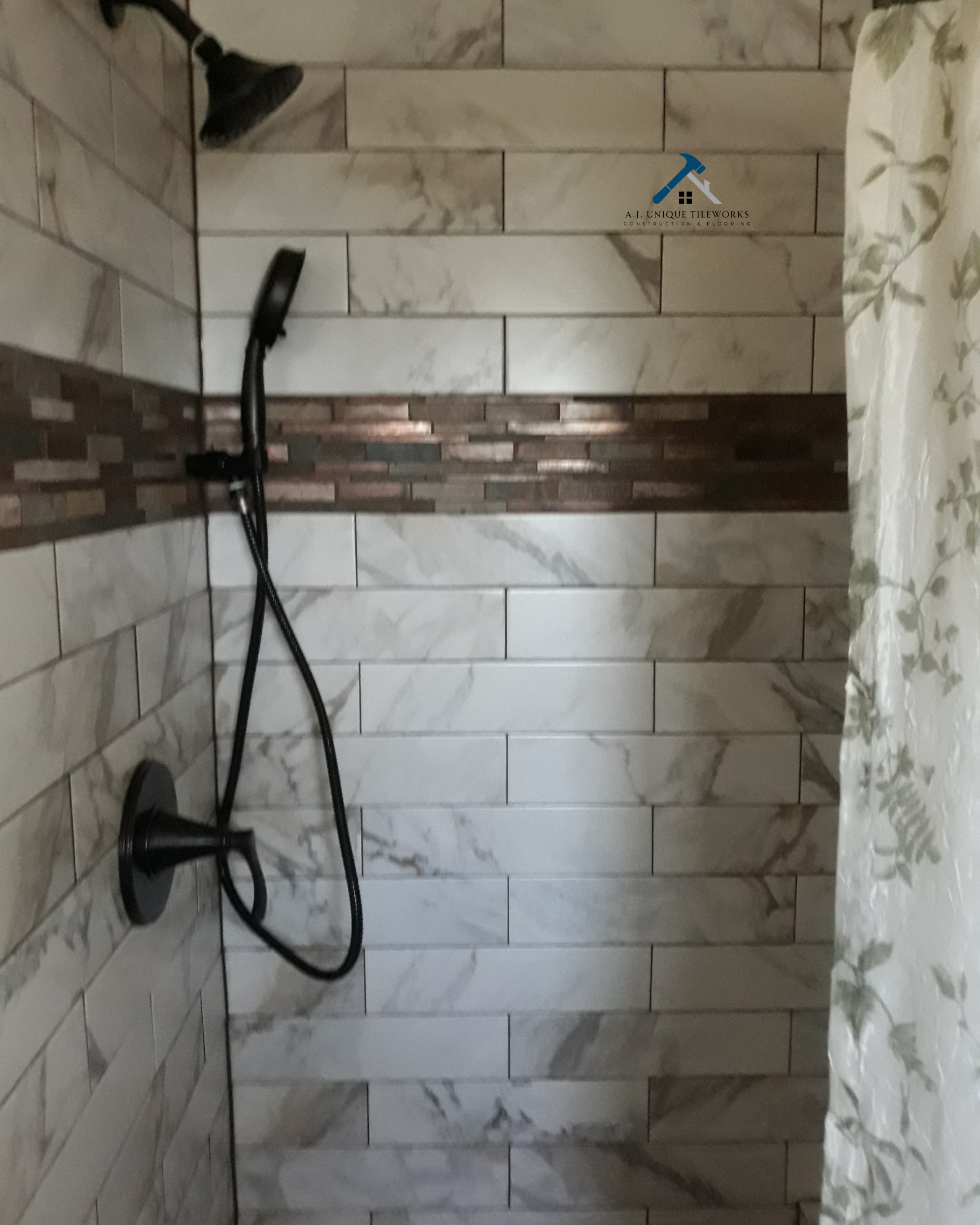 Bathroom shower with black showerhead and handheld shower, beige and brown tile walls, and a floral curtain on the right.