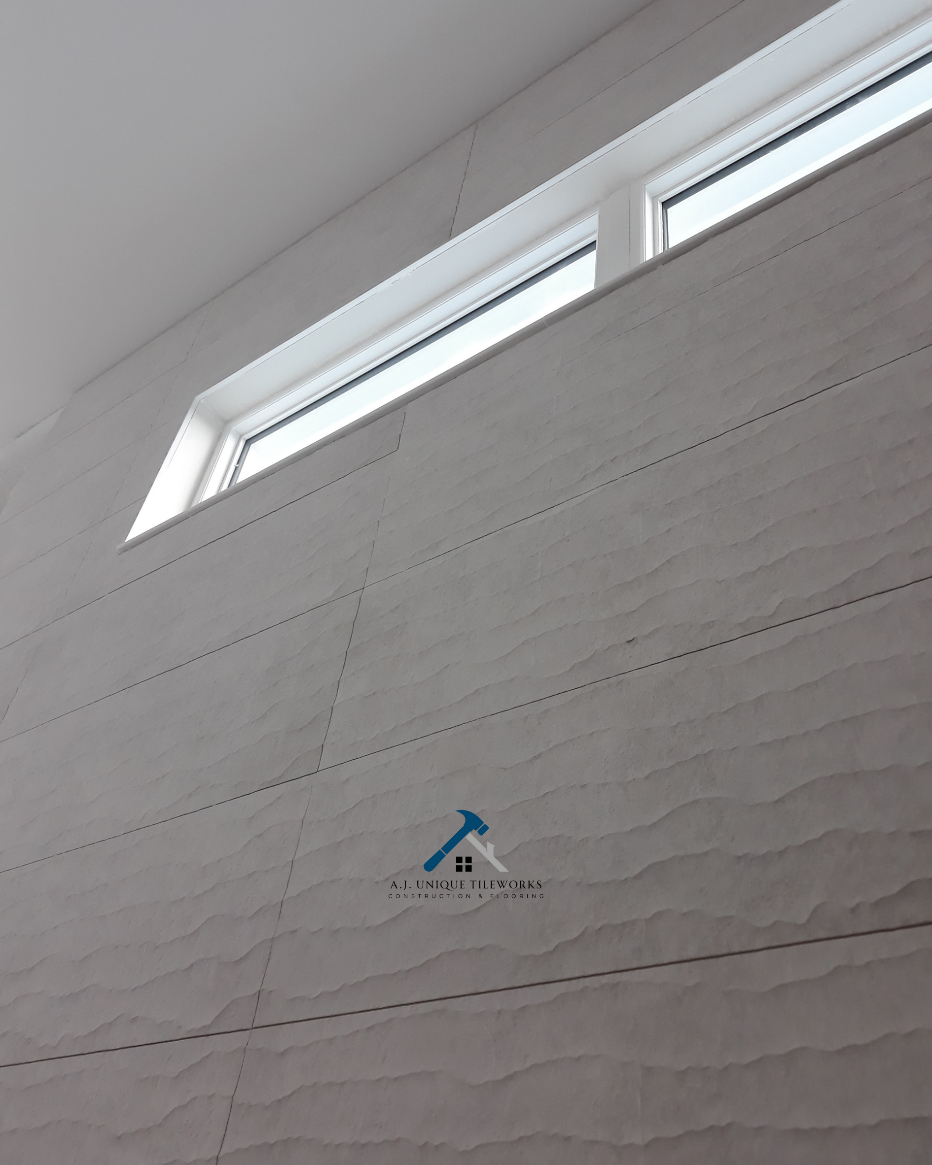 Interior wall with textured tile and a high horizontal window near the ceiling, with a logo of a house and hammer reading 'A.J. Unique Tileworks Construction & Flooring'.