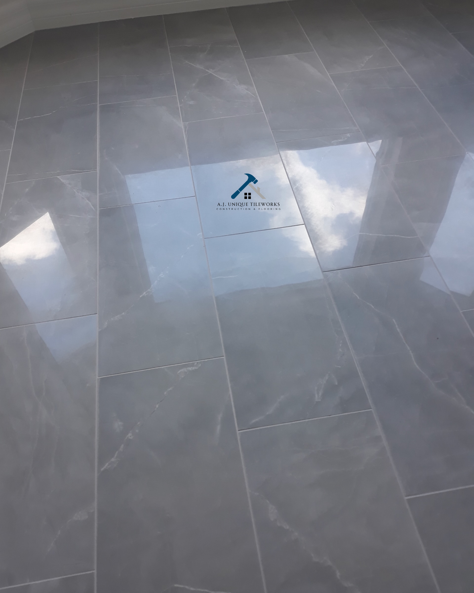 Shiny gray marble tile flooring with a company logo in the center featuring a hammer and house icon, along with the text 'A.J. Unique Tileworks Construction & Flooring'.