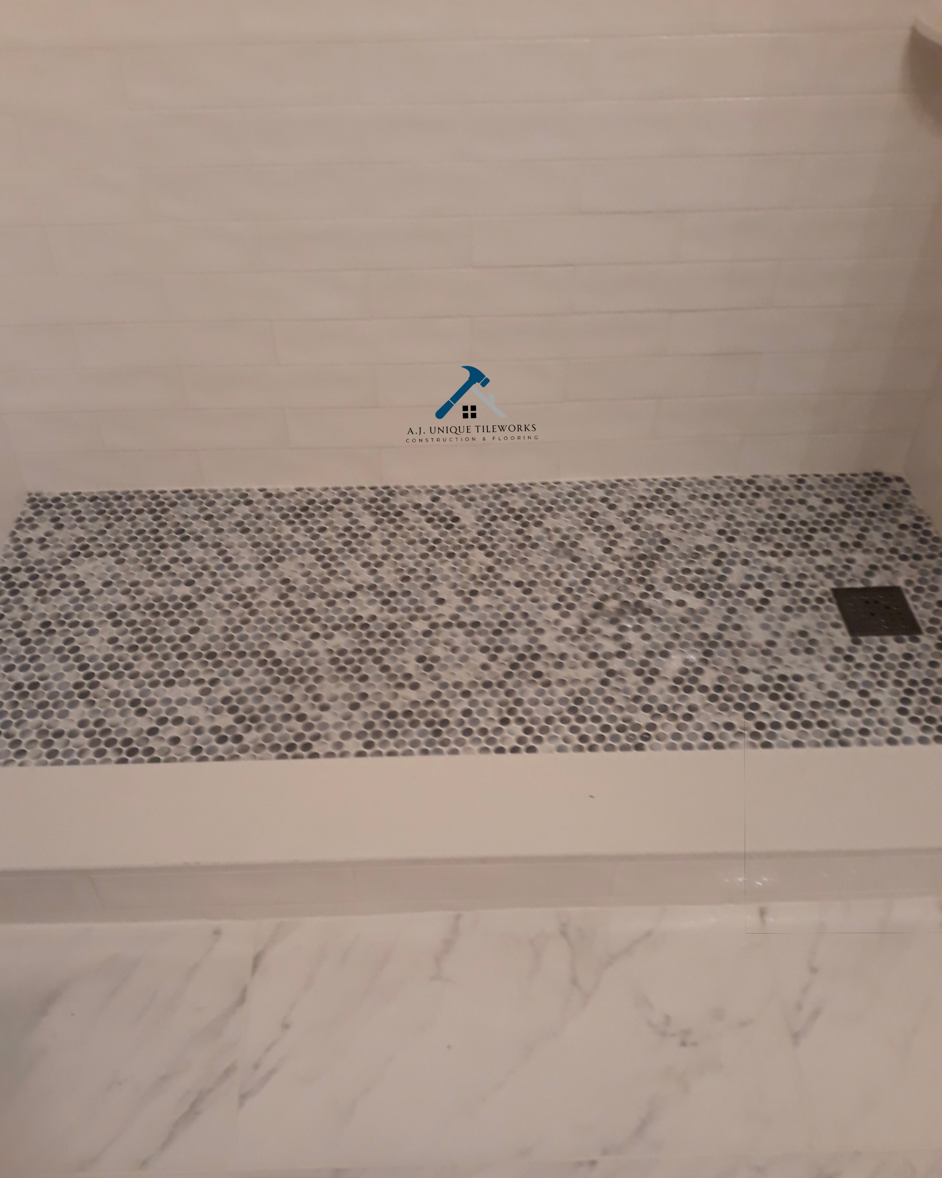 Bathroom shower area with small pebble mosaic tiles on the floor, white tile walls, a square drain at the floor, and a marble threshold at the entrance.