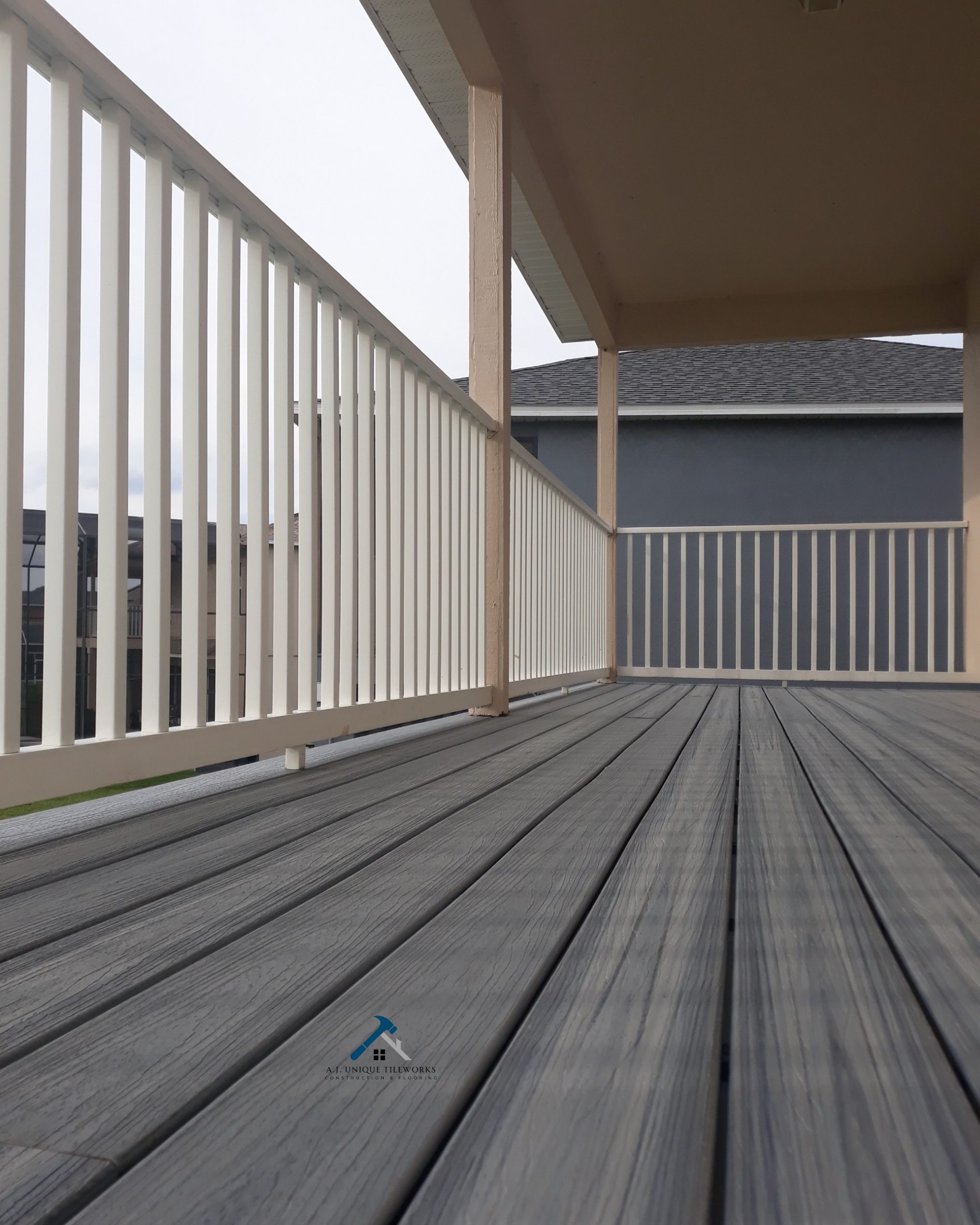 View of a porch with gray wooden decking and white railing, with neighboring houses visible in the background under an overcast sky.