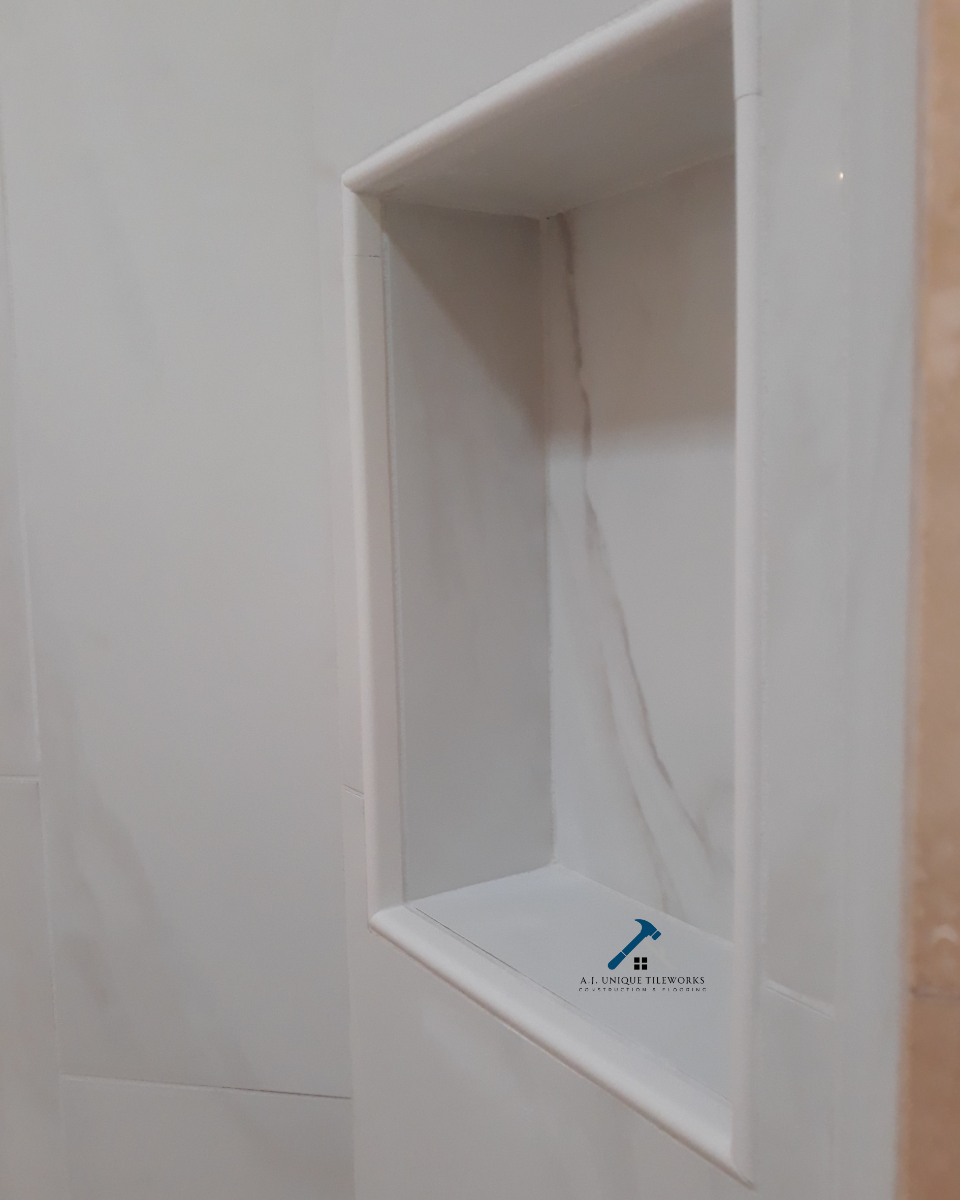 Close-up of a small recessed wall shelf with marble-like interior and white tile surrounding it, featuring a logo for A.J. Unique Tileworks.