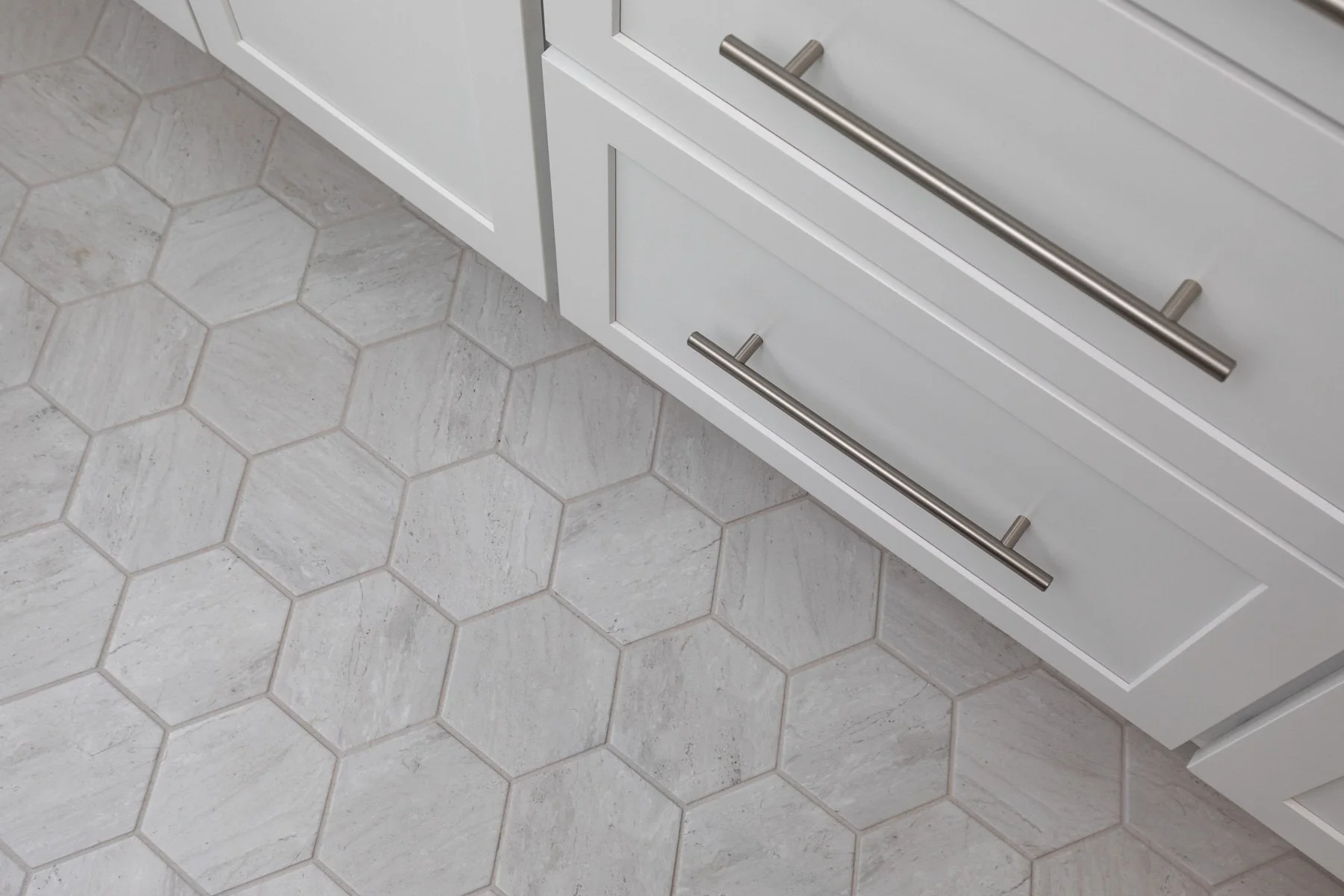 Close-up of a white kitchen cabinet with metal handles, showing a hexagonal tiled kitchen floor in light gray tones.