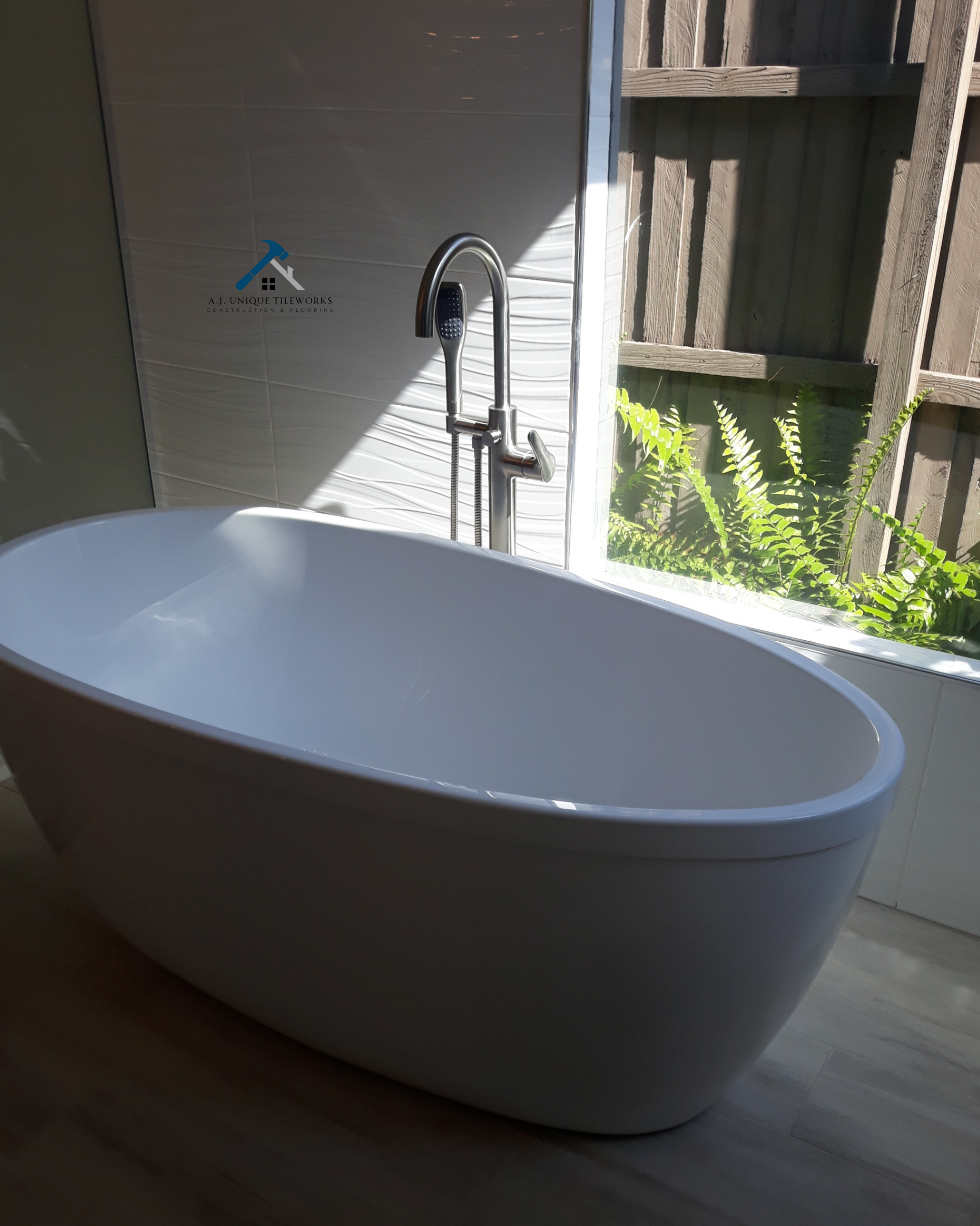 White freestanding bathtub next to a large window with a wooden fence and green plants outside. Modern chrome faucet with a handheld shower attachment. There is a logo on the wall behind the tub that reads 'A.J. Unique Tileworks Construction & Floori
