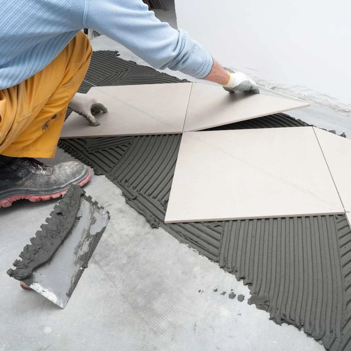 A worker installing large ceramic tiles on a floor with cement mortar and tile spacers for alignment.
