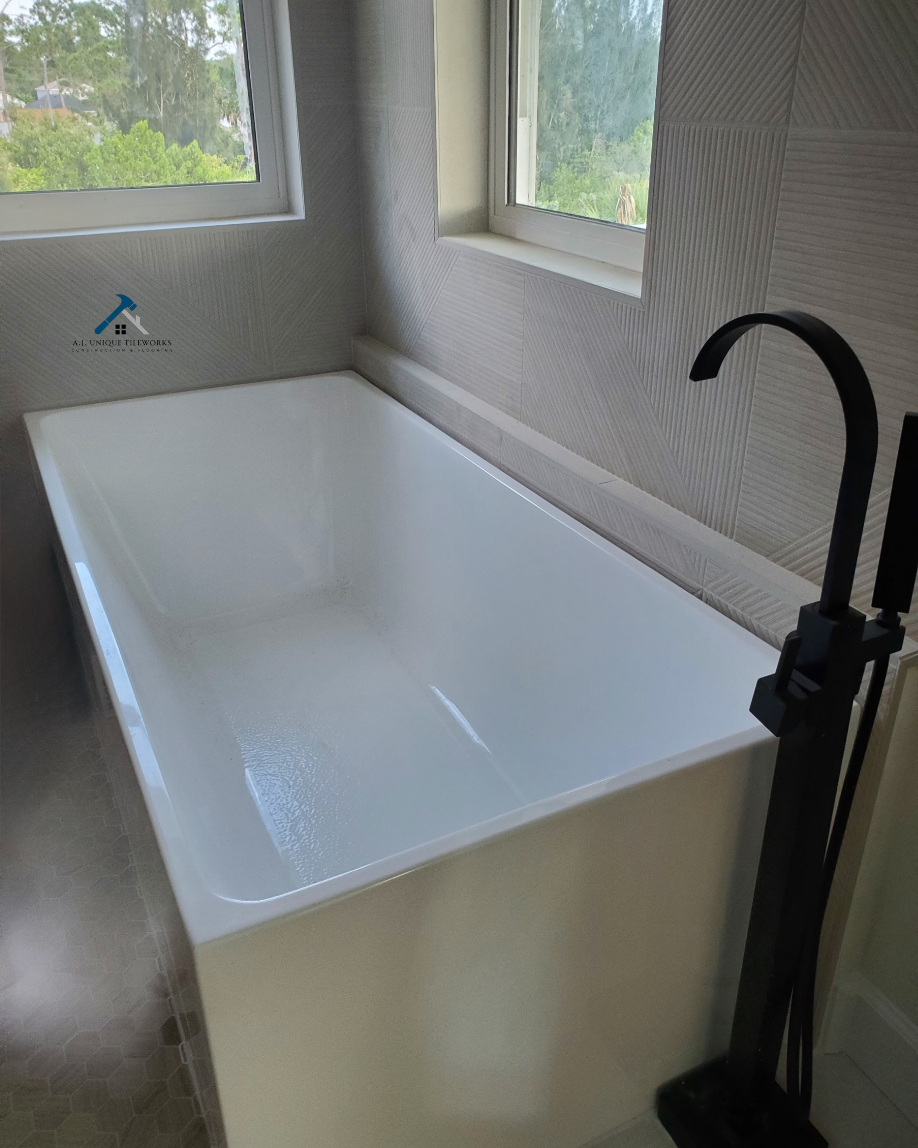 Empty white bathtub near two windows with greenery outside, black floor-mounted faucet at the corner of the bathtub, and textured gray walls.