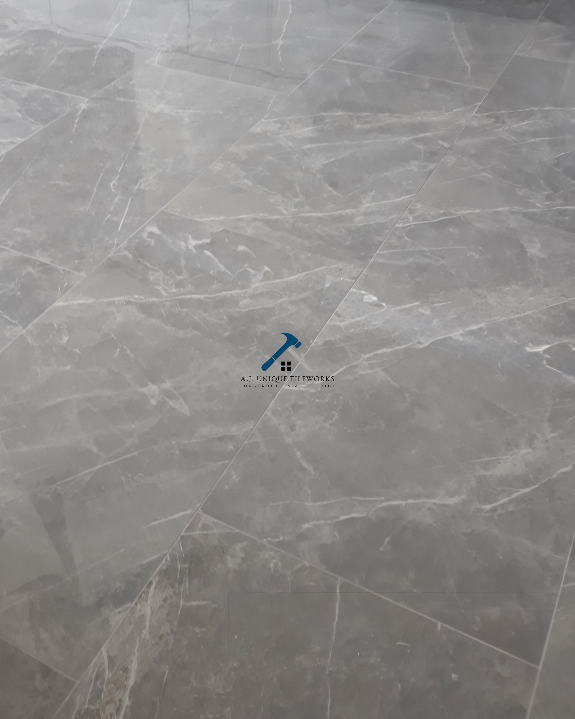Gray marble tile flooring with white veining and a logo in the center that shows a hammer, a house, and the text 'A.J. Unique Tileworks Construction & Flooring'.