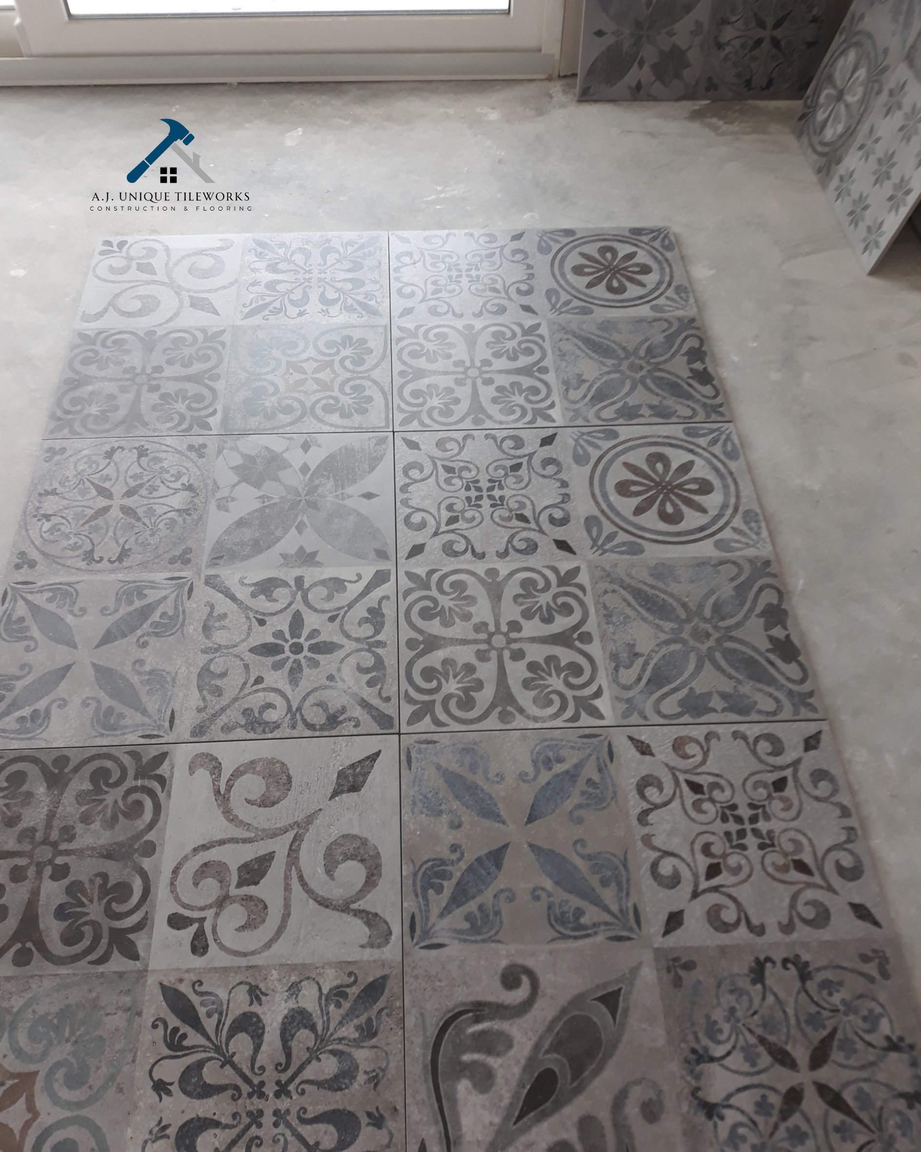 A floor with decorative gray and beige patterned tiles arranged in a grid, with a window or door at the back and some construction materials on the side.