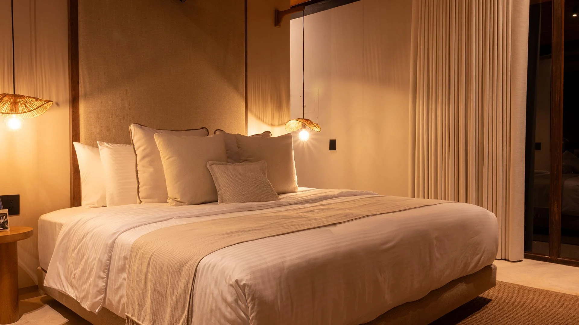 Cama doble con varias almohadas y edredón en una habitación luminosa con cortinas blancas y lámparas de techo decorativas.