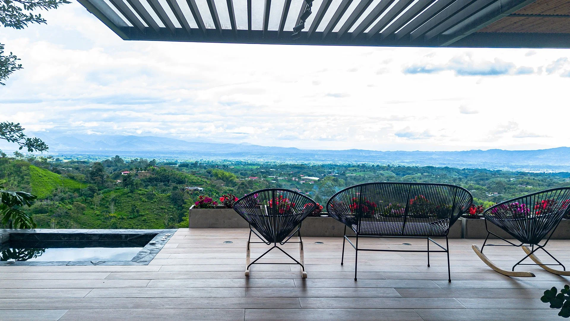Vista panorámica desde una terraza con muebles de mimbre negro y una pequeña piscina, rodeada de plantas y flores, con un paisaje de montañas y campo verde al fondo.