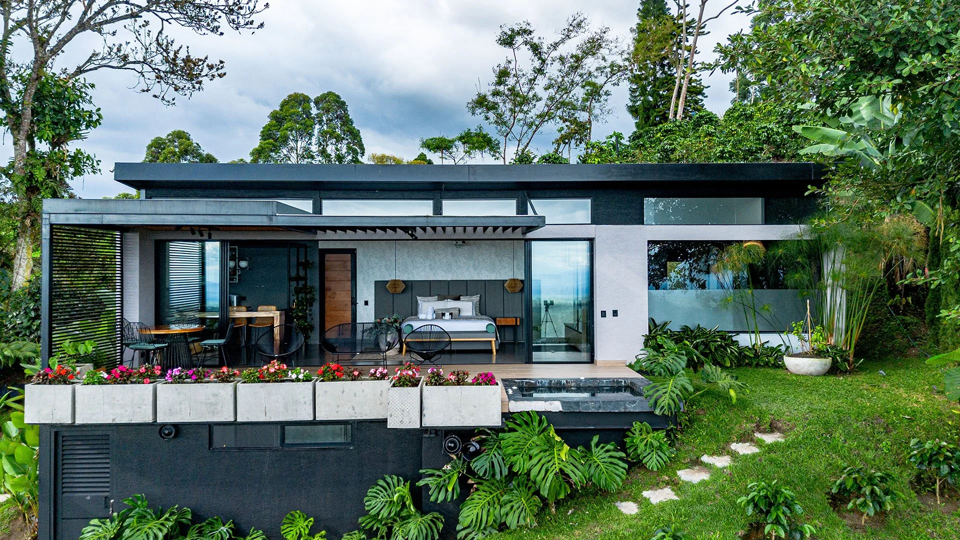Casa moderna en medio de un jardín con numerosas plantas y flores, con área de descanso y ventanas grandes en un entorno natural.