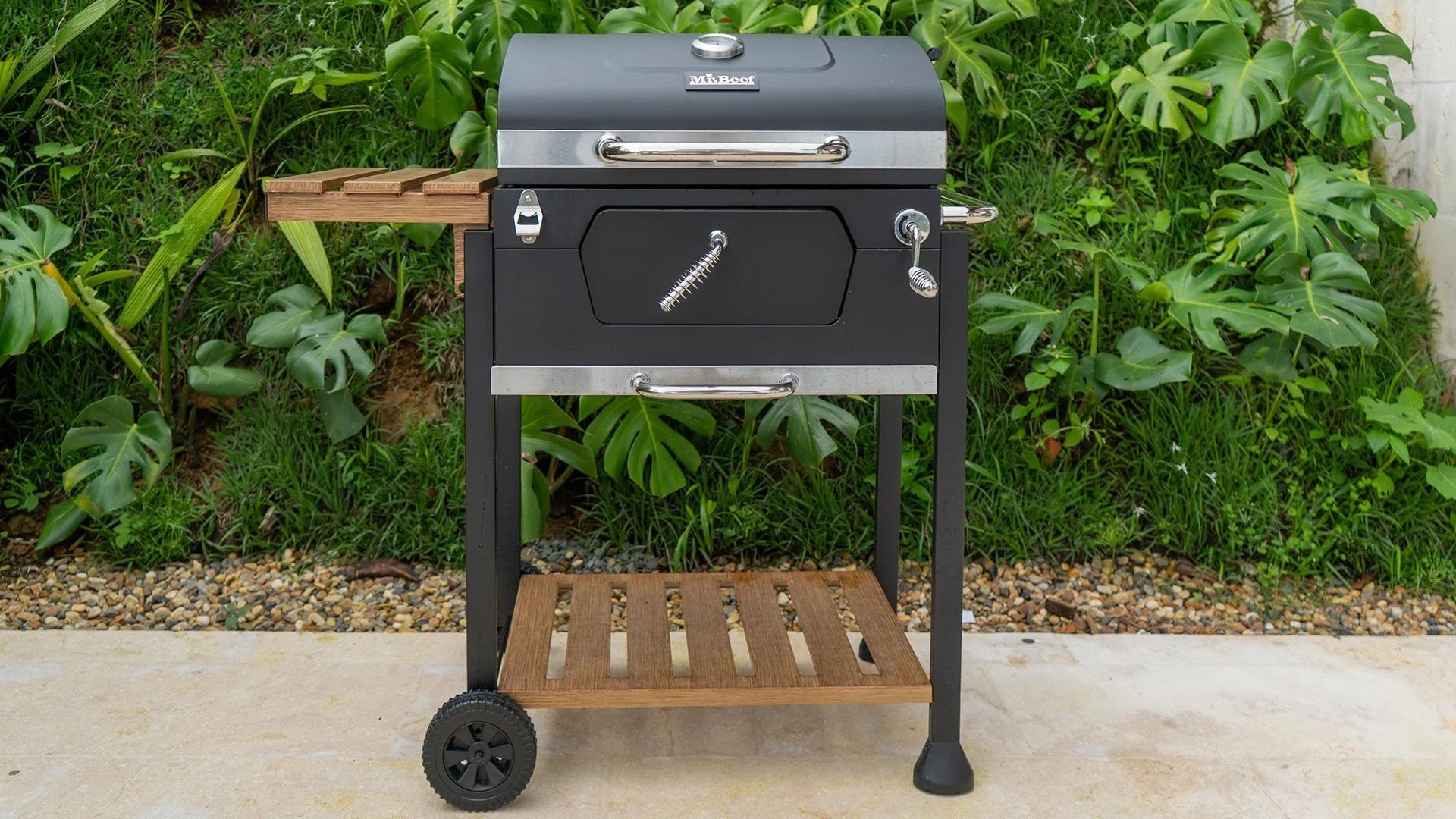 Parrilla de carbón negra en un patio con plantas verdes de fondo. Tiene ruedas y una bandeja de madera a la derecha.