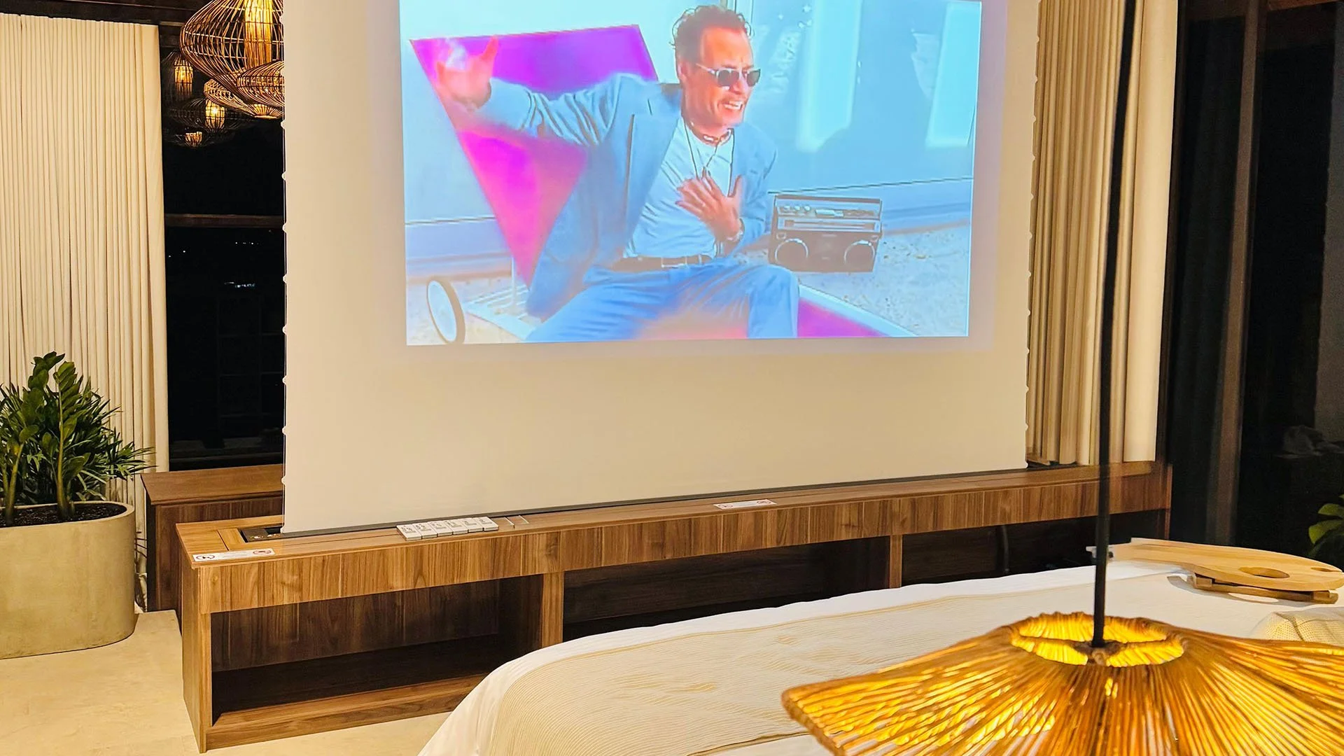 Pantalla de proyector en una habitación, mostrando la imagen de un hombre con gafas de sol y ropa azul, sentado en una silla con una mano en el pecho y la otra levantada, con una caja de stereo y un fondo colorido de tonos rosados y azules.