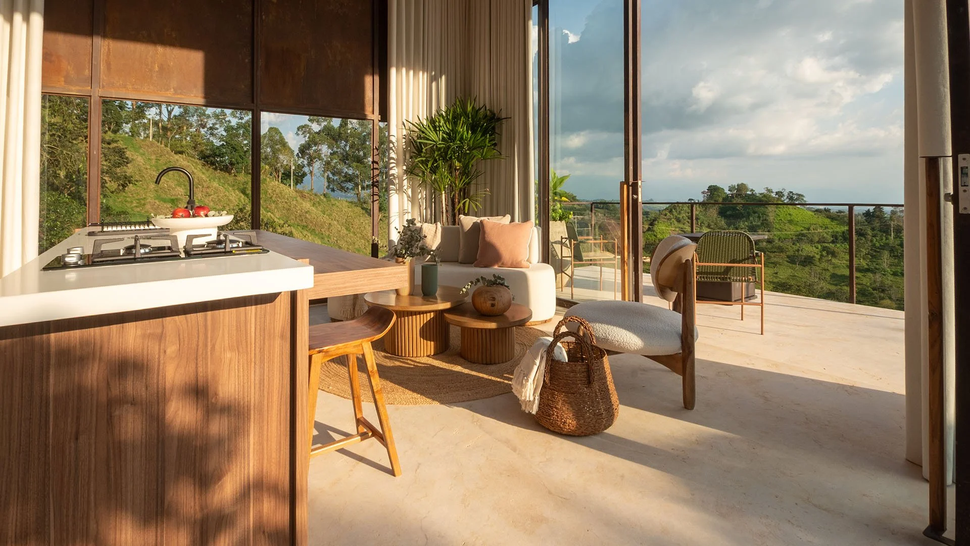 Sala de estar y balcón con vista a la naturaleza, muebles de madera, plantas y ventanas grandes que dejan entrar la luz.
