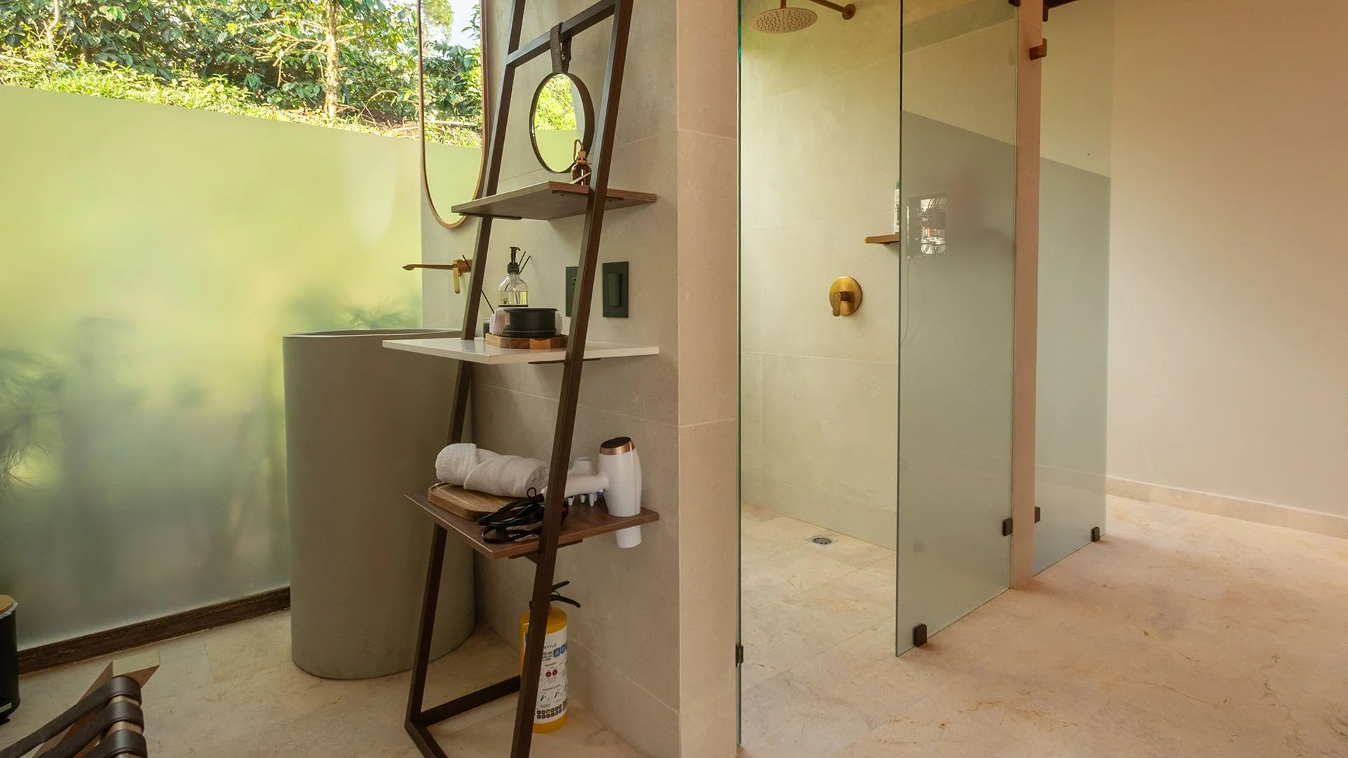 Vista de baño con paredes de vidrio, ducha con ducha de lluvia, estantería de madera con accesorios y un espacio abierto con ventanas de vidrio que dejan ver vegetación exterior.