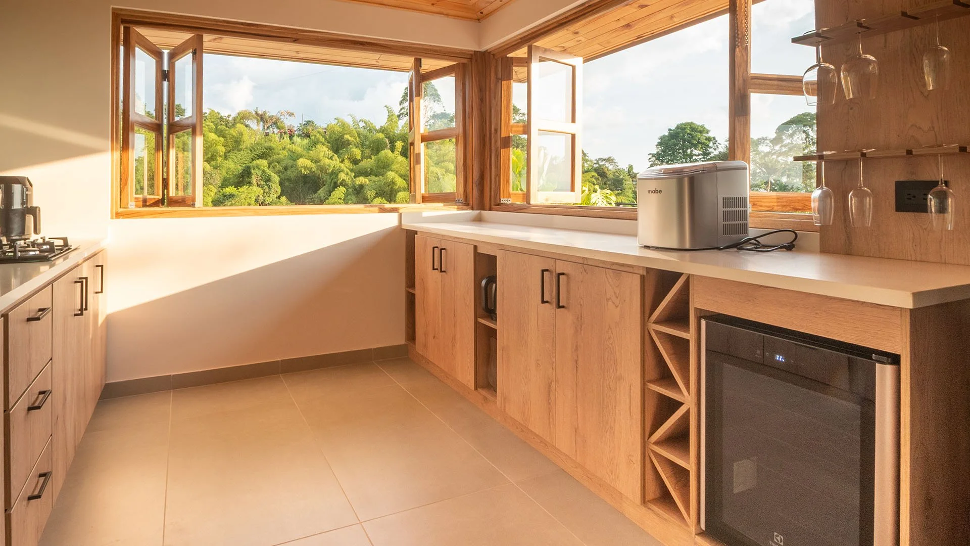 Cocina con ventanas que muestran un paisaje verde y cielo azul, gabinetes de madera, electrodomésticos y copas de vino colgando.