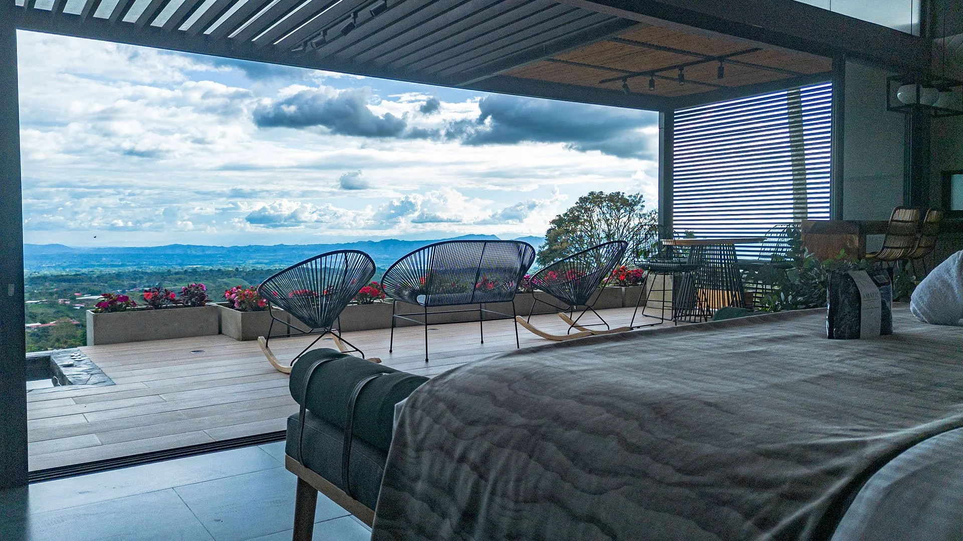 Vista desde habitación con cama y balcón con sillas y vista panorámica de montañas y cielo nublado.