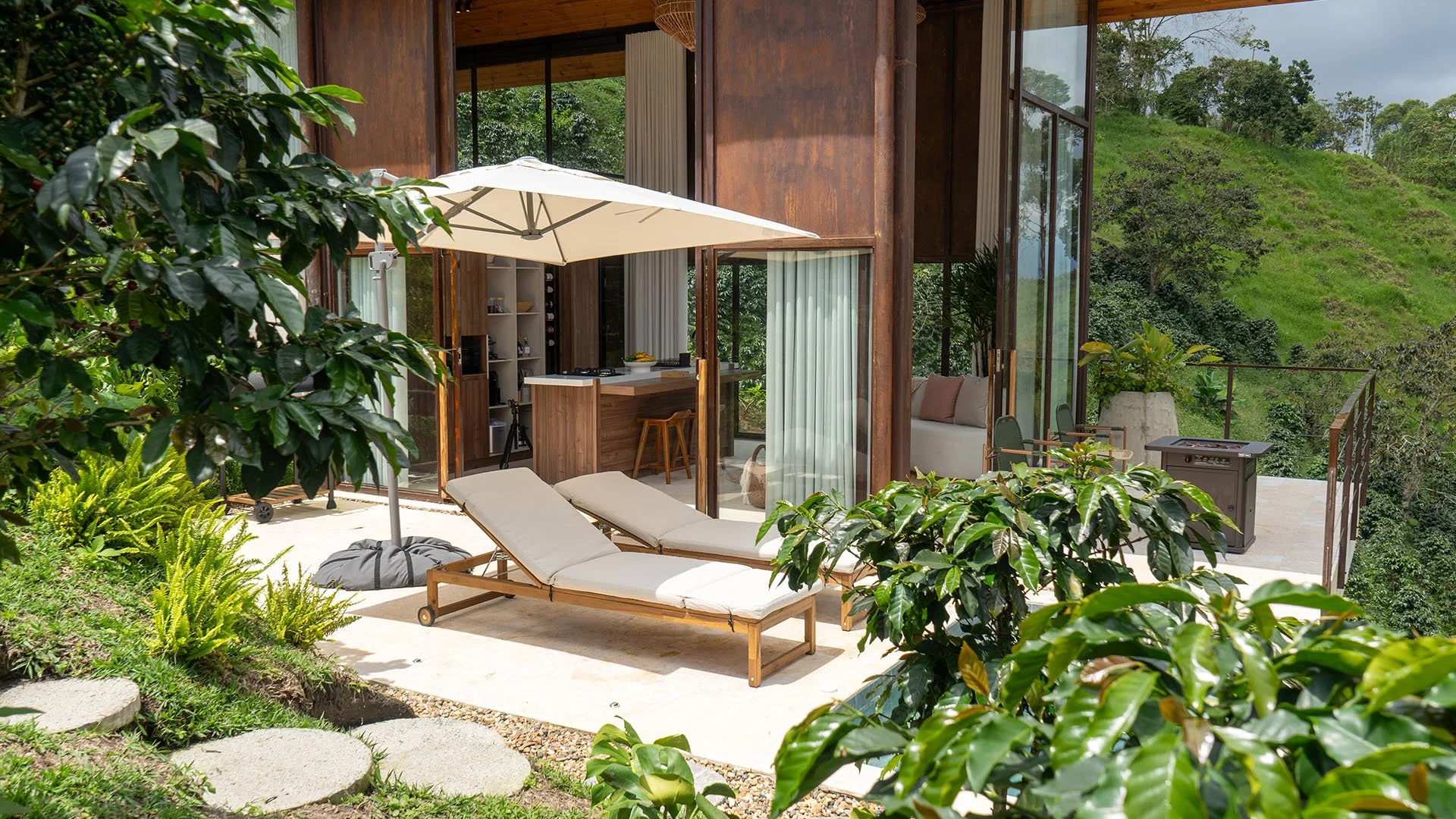 Terraza con tumbonas bajo un paraguas, rodeada de vegetación y vista a colinas verdes en un entorno natural.