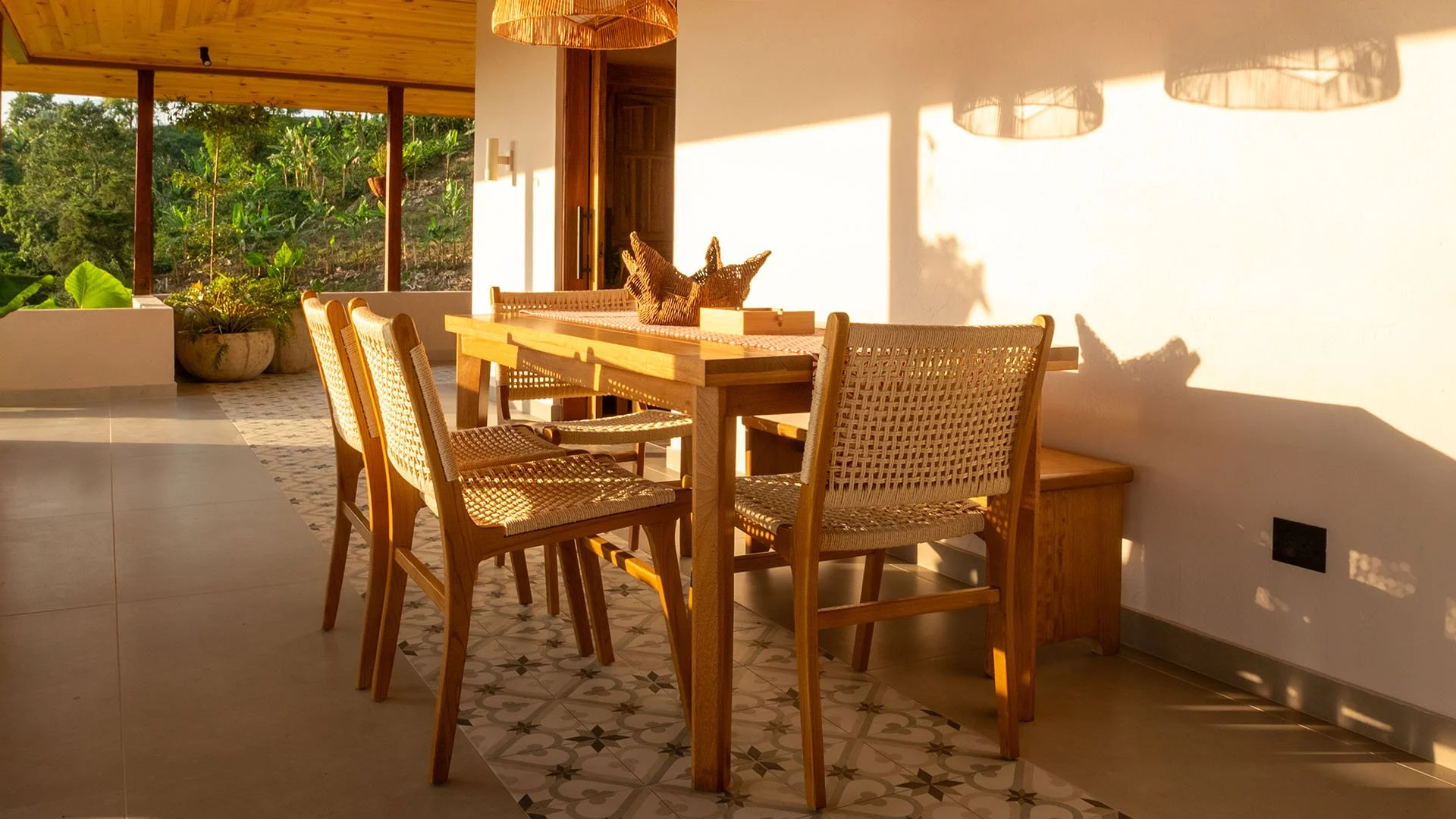 Sala de comedor con mesa de madera y sillas de mimbre, iluminada por luz cálida y sombras del sol, con plantas verdes en el fondo y paredes blancas.