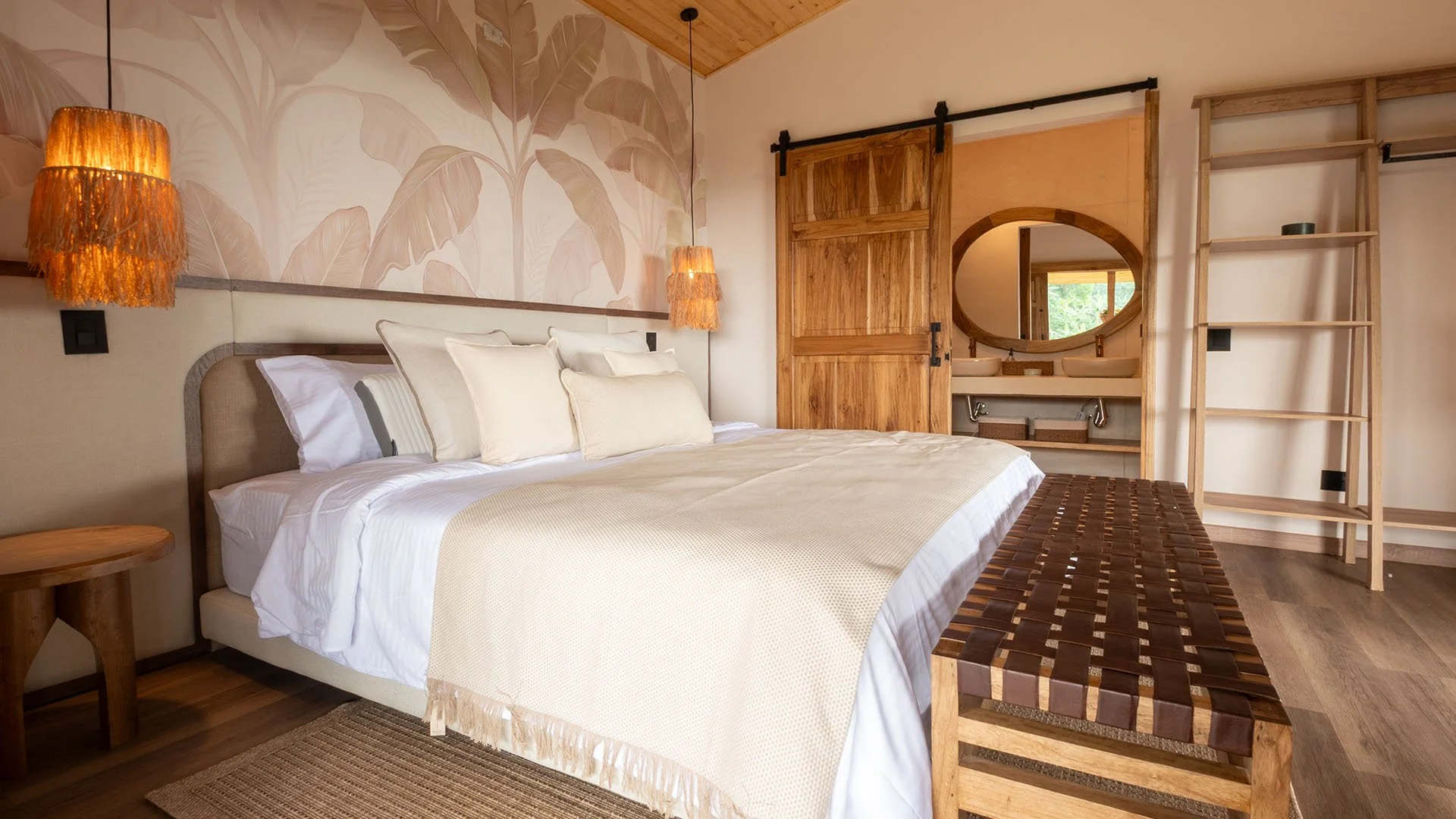Habitación con cama grande, paredes con estampado de hojas, lámparas de fibra natural, puertas de madera, espejo ovalado y estructura de madera para colgar ropa.