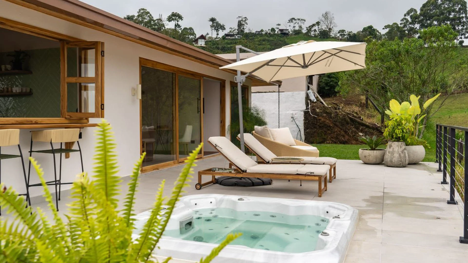 Terraza con tumbonas, sombrilla y jacuzzi, rodeada de plantas y con vista a colinas verdes.